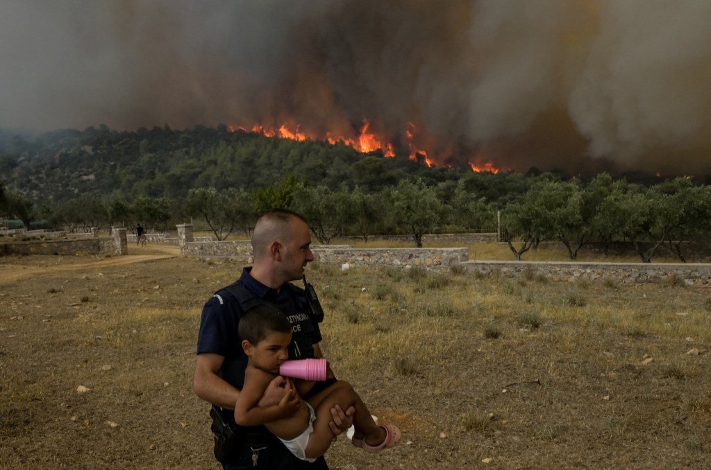 A policeman evacuates a child from wildfire at the village of Agios Charalabos, near Athens, on July 18, 2023. Europe braced for new high temperatures on July 18, 2023, under a relentless heatwave and wildfires that have scorched swathes of the Northern Hemisphere, forcing the evacuation of 1,200 children close to a Greek seaside resort. Health authorities have sounded alarms from North America to Europe and Asia, urging people to stay hydrated and shelter from the burning sun, in a stark reminder of the effects of global warming. (Photo by Aris MESSINIS / AFP)