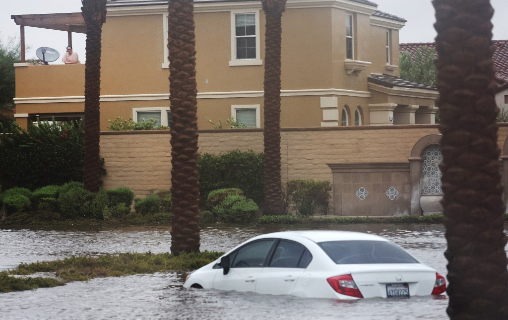 CATHEDRAL CITY, CALIFORNIA - AUGUST 20: A person looks on from a residence toward a car partially submerged in floodwaters as Tropical Storm Hilary moves through the area on August 20, 2023 in Cathedral City, California. Southern California is under a first-ever tropical storm warning as Hilary impacts parts of California, Arizona and Nevada. All California state beaches have been closed in San Diego and Orange counties in preparation for the impacts from the storm which was downgraded from hurricane status. Mario Tama/Getty Images/AFP (Photo by MARIO TAMA / GETTY IMAGES NORTH AMERICA / Getty Images via AFP)