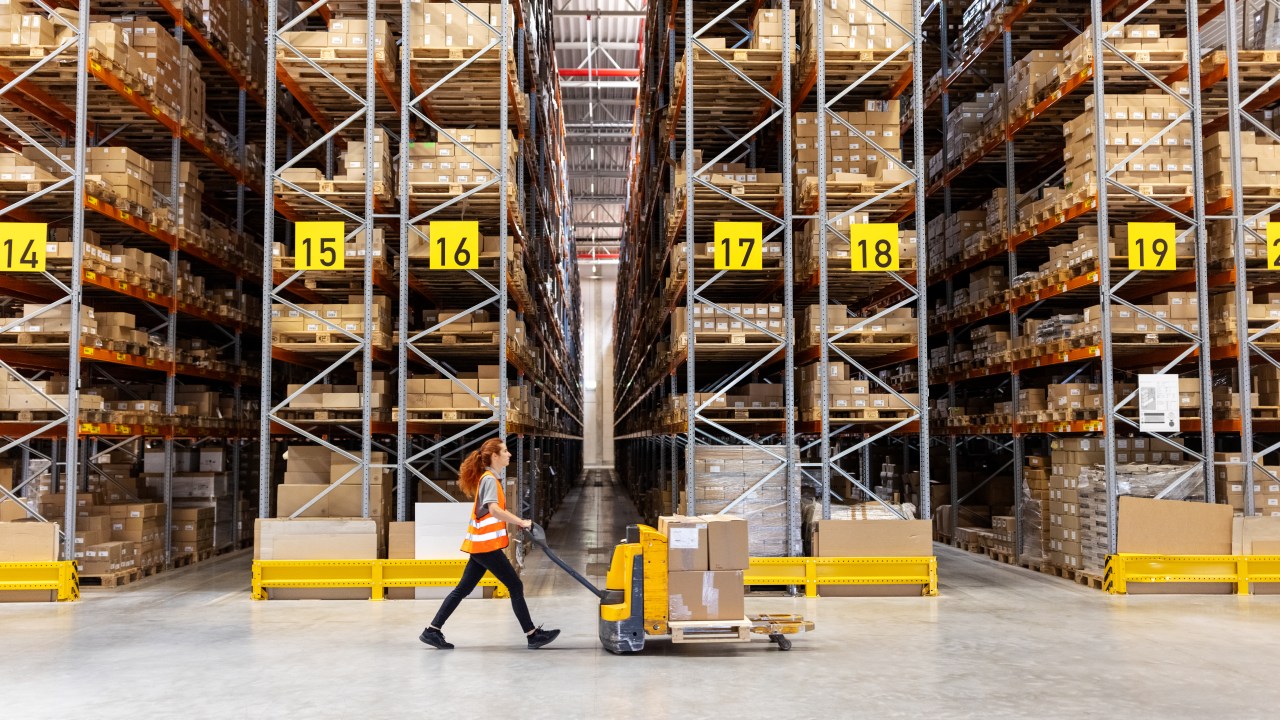 Side view of a female worker pushing hand truck with boxes in a storehouse. Warehouse employee walking with cardboard boxes on pallet jack along large racks.