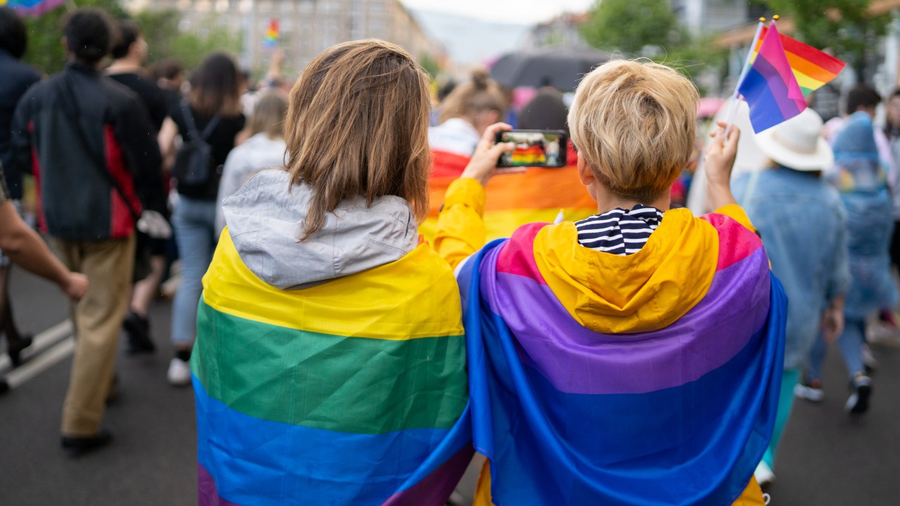 Duas mulheres enroladas em bandeiras LGBT seguram bandeiras do movimento Bi durante parada LGBT