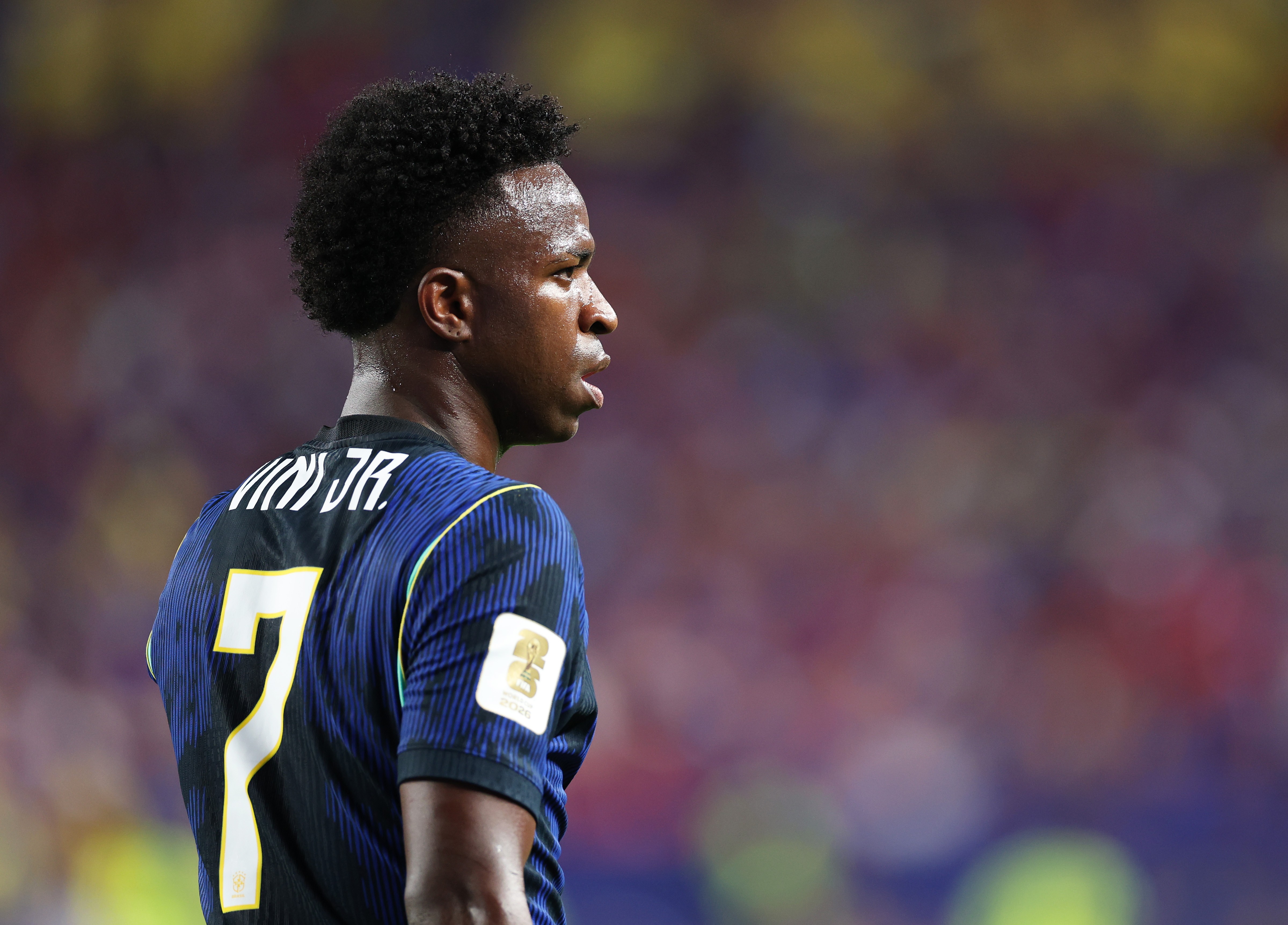Jogador de futebol negro, com cabelo crespo curto, vestindo camisa azul escura com listras finas e o número 7 amarelo nas costas, olhando para a direita com expressão séria, em campo de futebol com público desfocado ao fundo