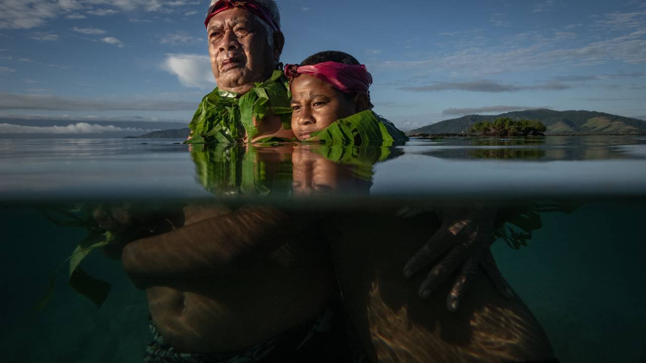 Lutando, Não Afundando: Lotomau Fiafia (72), um ancião da comunidade, se posiciona com seu neto John no local onde ele lembra que a linha costeira costumava estar quando era criança. Baía de Salia, Ilha Kioa, Fiji, 8 de agosto de 2023 A comunidade de 500 pessoas na Ilha Kioa cresceu a partir de um assentamento de pessoas que buscaram refúgio na década de 1940 devido ao aumento do nível do mar em Tuvalu, uma ilha ao norte.