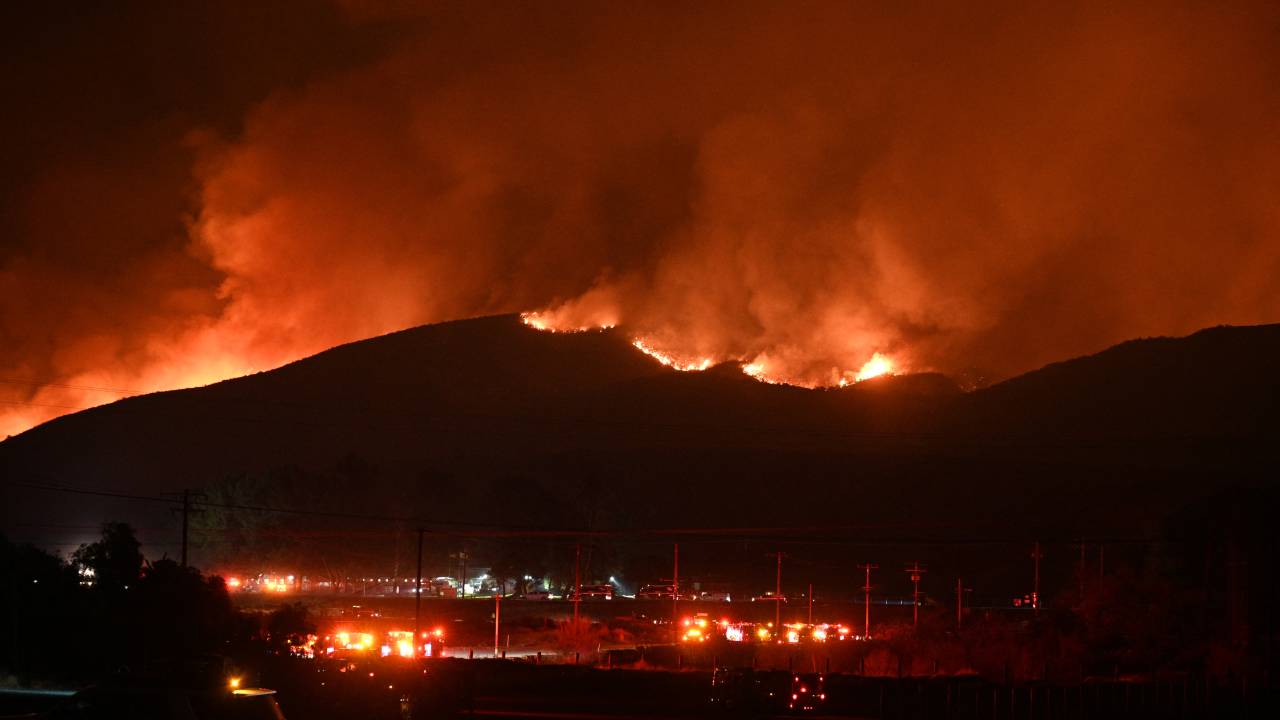 Fire trucks and public security vehicles are seen in the foreground as red glowing flames and smoke rise from the Hughes Fire in Castaic in northwest Los Angeles County, California, on January 22, 2025. A new wildfire erupted north of Los Angeles on January 22, exploding in size and sparking thousands of evacuation orders in a region already staggering from the effects of huge blazes. Ferocious flames were devouring hillsides near Castaic Lake, spreading rapidly to cover 5,000 acres (2,000 hectares) in just over two hours. (Photo by Robyn Beck / AFP)