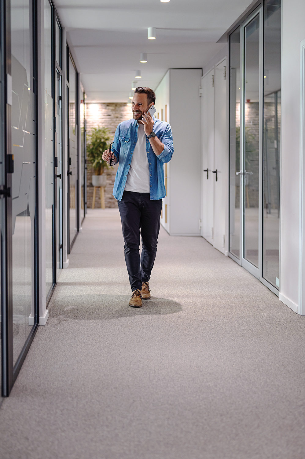 Full length of male manager holding glasses and discussing ideas over phone call in office corridor VALE TUDO - Andar ao telefone: medida simples quebra longos períodos de inatividade