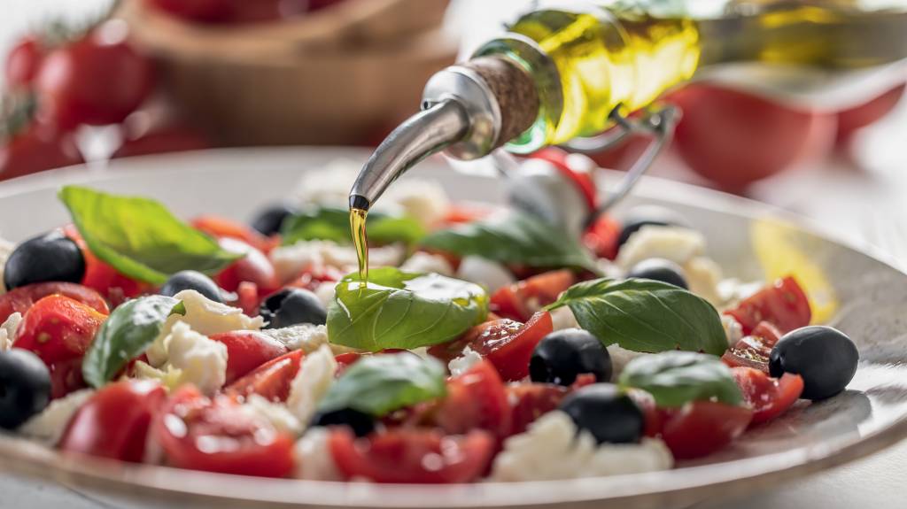 Close-up of pouring olive oil into an Italian Caprese salad. Mediterranean vegetarian appetizer with olive oil. VELHO SEGREDO - Dieta mediterrânea: com enfoque em vegetais e proteínas magras, já foi associada a controle do peso