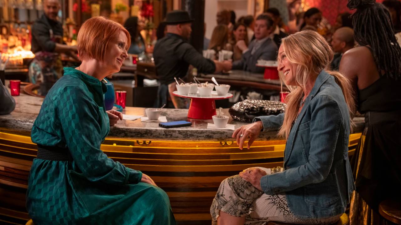 Duas mulheres sentadas em um bar, uma com cabelo ruivo usando um vestido verde e a outra com cabelo loiro usando uma jaqueta jeans e uma saia estampadas conversando e sorrindo. Há outros clientes e funcionários ao fundo, e o bar está iluminado com luzes douradas.