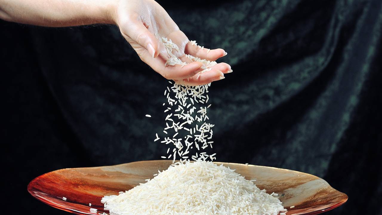 Grains of white rice falling through outstretched fingers into shallow glass bowl.