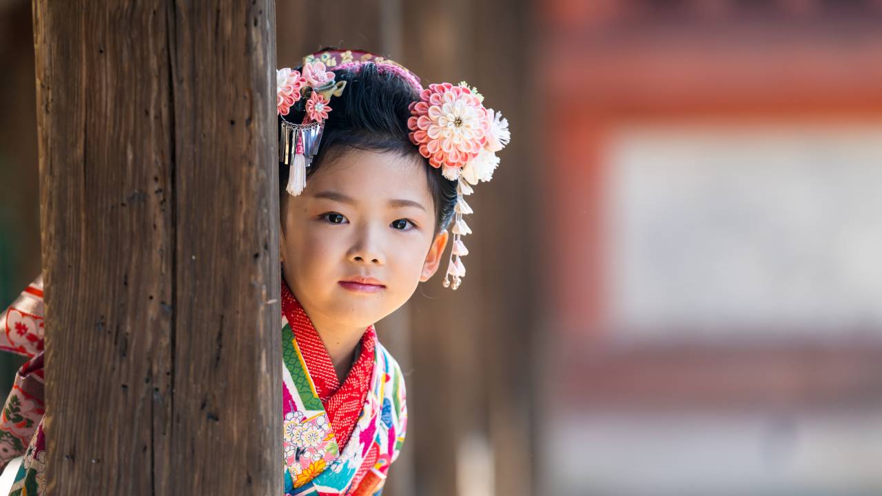 A young Japanese girl wearing a traditional kimono peeking out from behind a wooden post