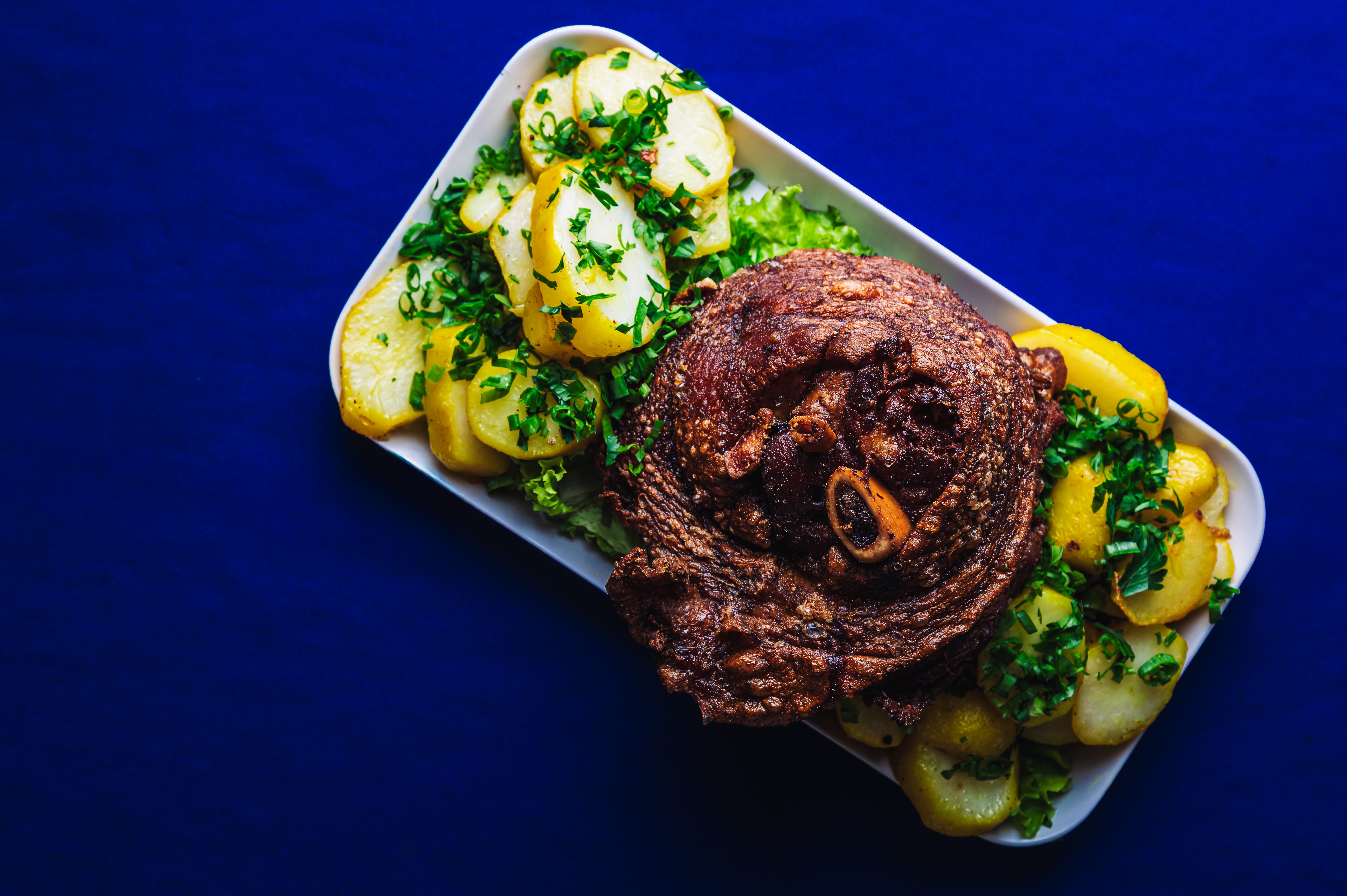 Prato branco com joelho de porco assado, dourado e crocante, acompanhado de batatas cozidas e fatiadas, salpicadas com cheiro-verde picado sobre folhas de alface, em fundo azul escuro