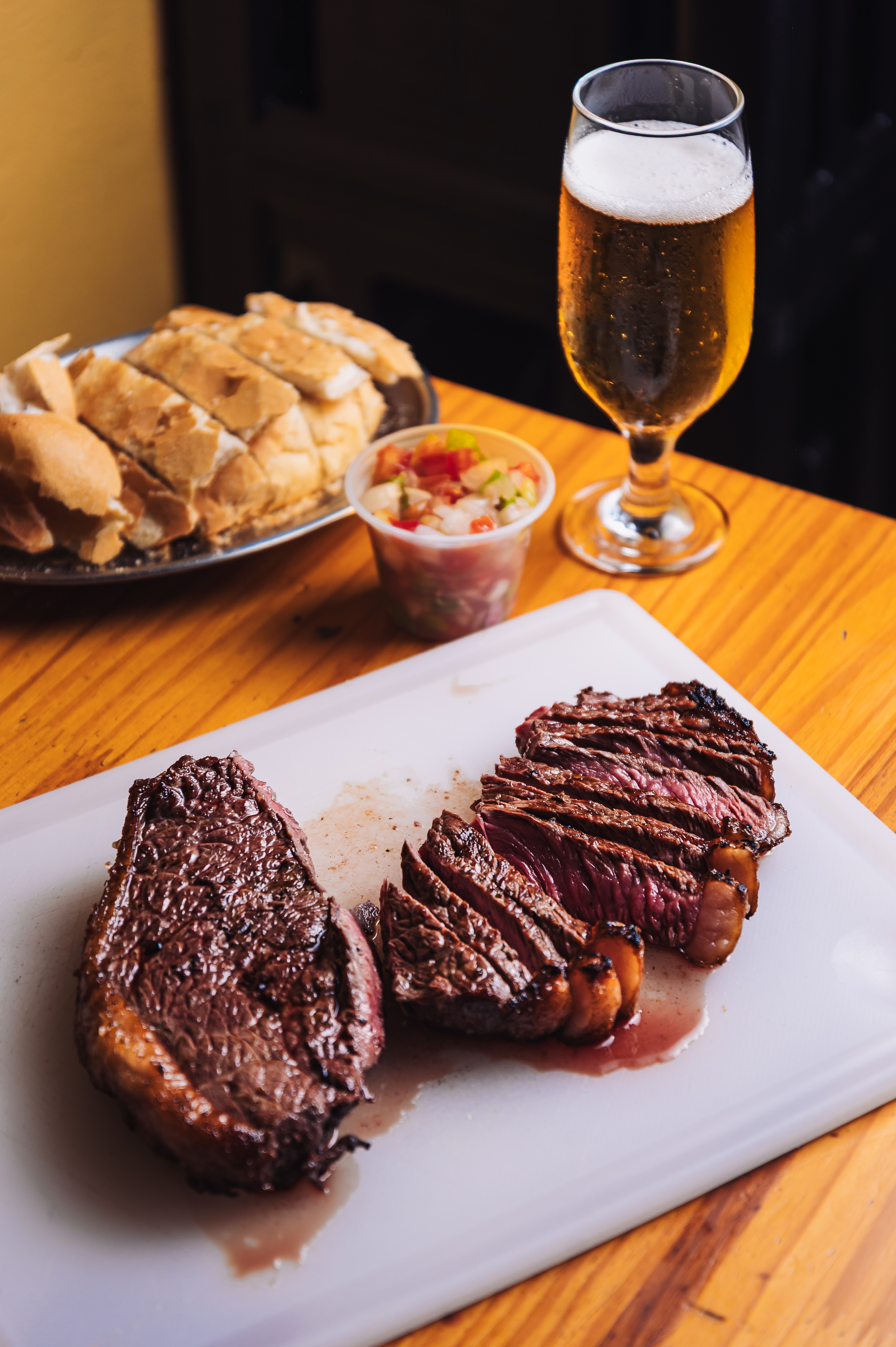 Picanha grelhada, uma pe&ccedil;a inteira e outra fatiada, sobre t&aacute;bua branca. Ao fundo, p&atilde;o fatiado, vinagrete em pote e um copo de cerveja clara com espuma, tudo sobre mesa de madeira.