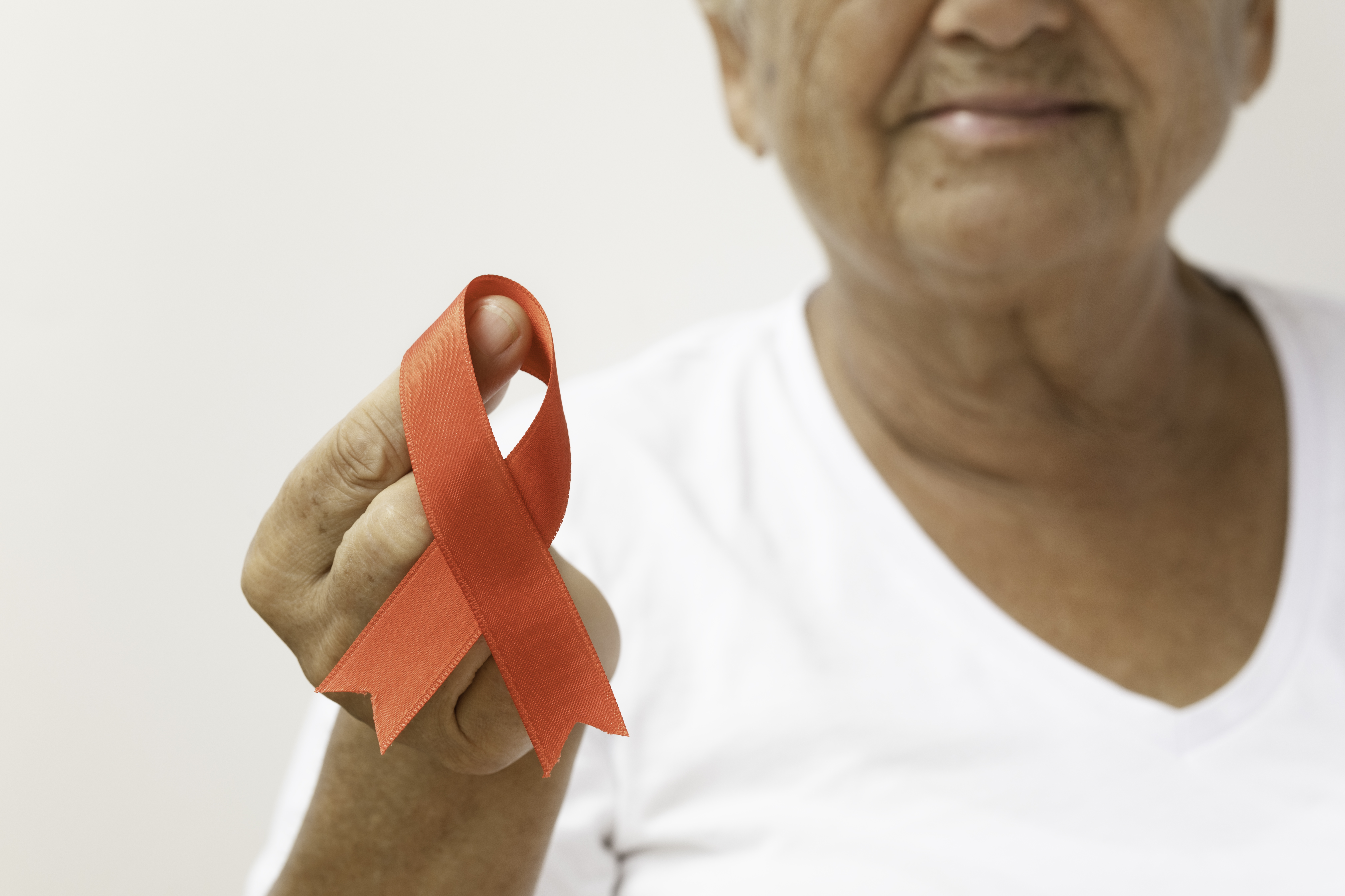 Senior woman is holding red awareness ribbon in front of white background. Some issues for what the red awareness ribbon stands for are AIDS, addiction, alcohol dependence, blood Cancer, heart defects, drug addiction, red ribbon week, Hypertension, Tuberculosis and Zika Virus.