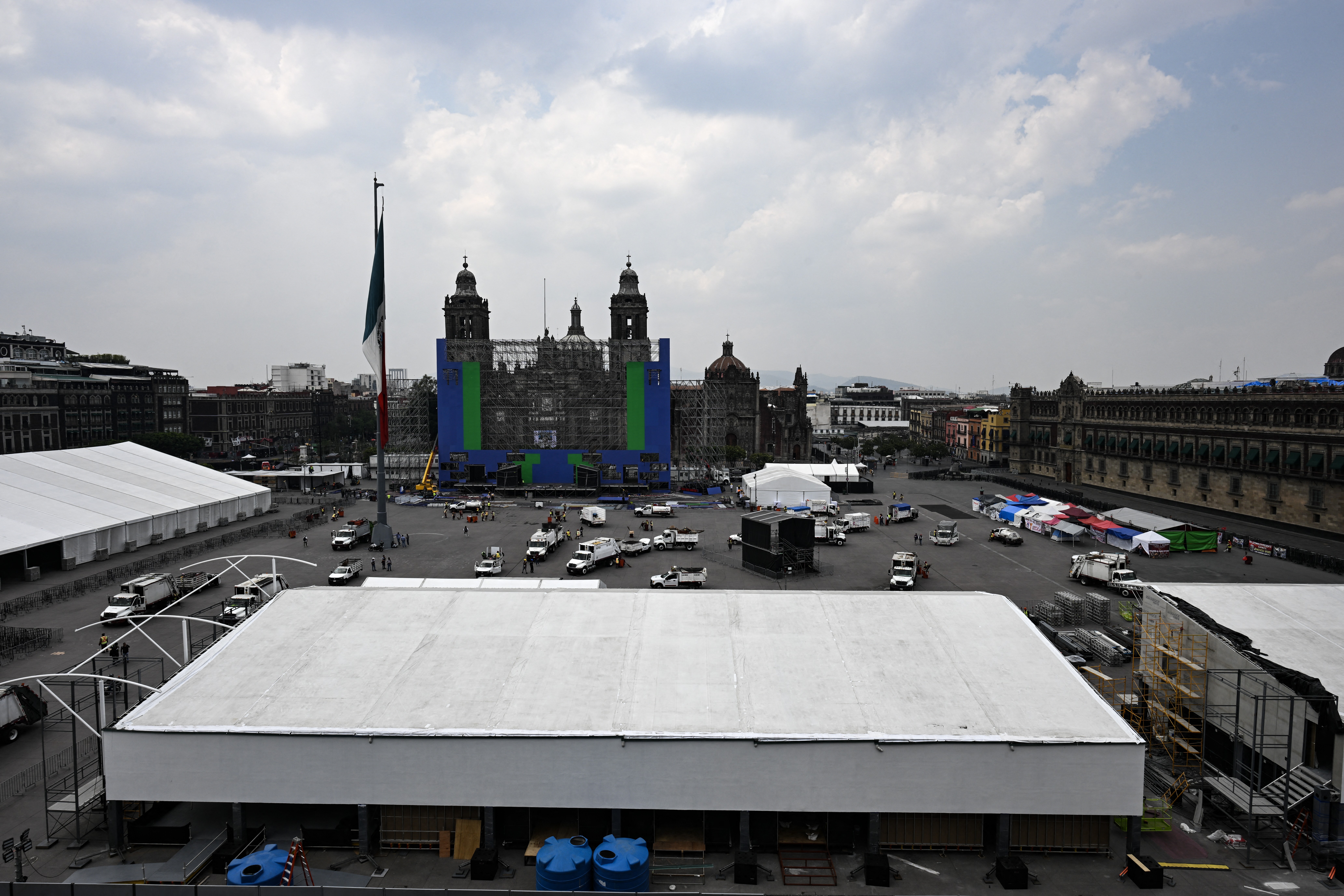 Praça Zócalo, Cidade do México, com o Palácio Nacional ao fundo, parcialmente coberto por lonas azuis e verdes. Tendas brancas e veículos de serviço ocupam a praça, sob um céu nublado. Uma bandeira mexicana tremula à esquerda
