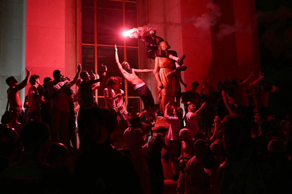 Multidão em protesto noturno, iluminada por luz vermelha. Um homem sem camisa segura um sinalizador, enquanto outro está sobre os ombros de uma estátua. Fumaça vermelha sobe, e pessoas ao redor levantam os braços em celebração ou protesto