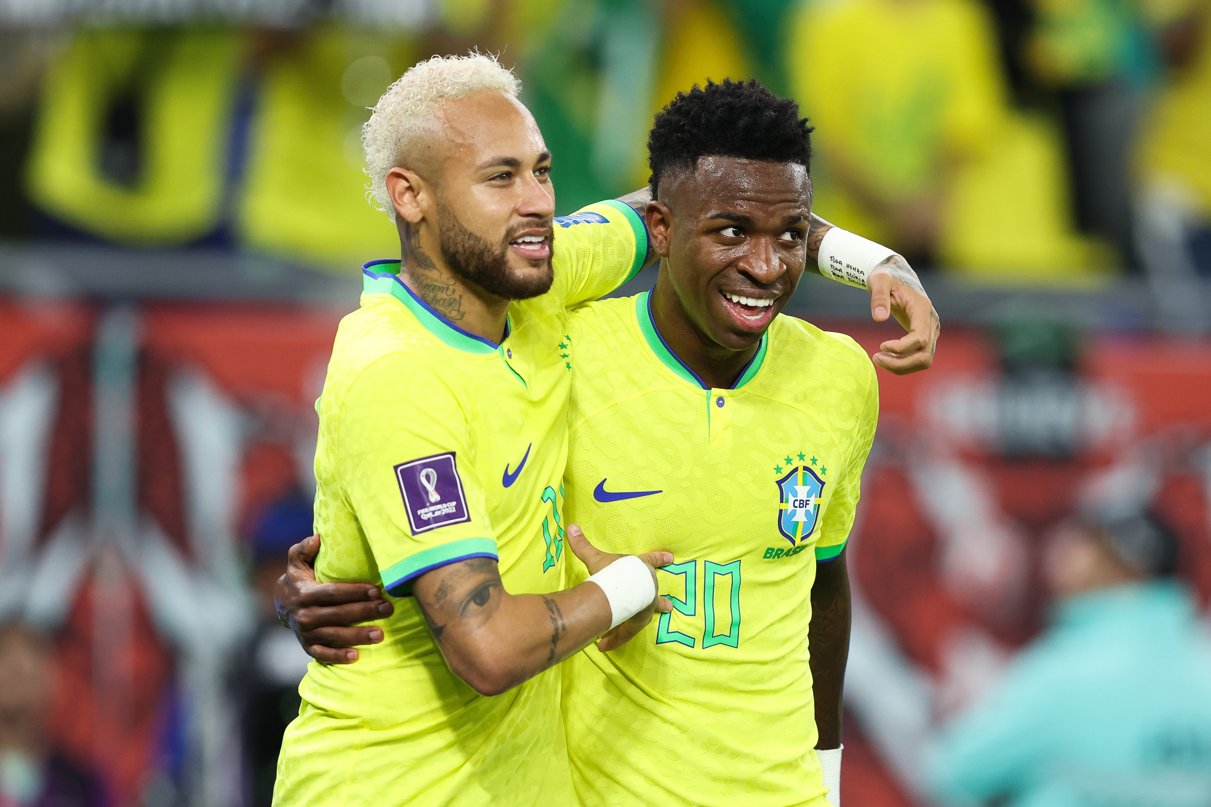 Neymar, com cabelo loiro e barba, abraça Vinicius Jr, que sorri, ambos vestindo camisas amarelas da seleção brasileira de futebol, em campo
