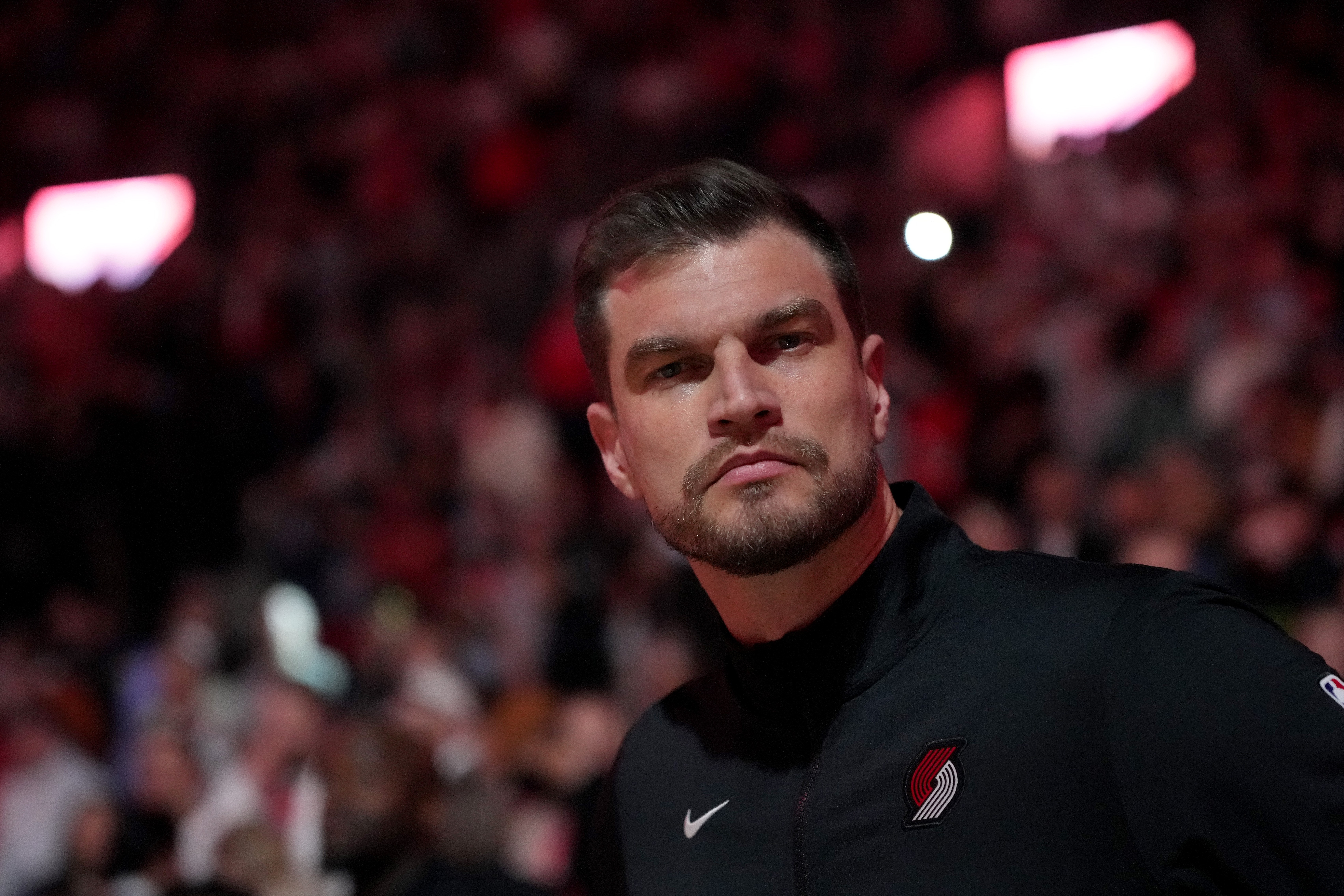 Homem branco de barba e cabelo escuro, vestindo jaqueta preta com logo do Portland Trail Blazers, olhando sério para frente, com público desfocado ao fundo em ambiente escuro e iluminado por luzes vermelhas