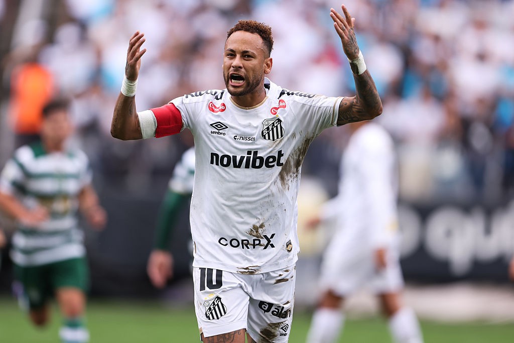Neymar Jr. em campo, vestindo camisa branca do Santos suja de terra, com braços erguidos e boca aberta, expressando frustração. Outros jogadores desfocados ao fundo.
