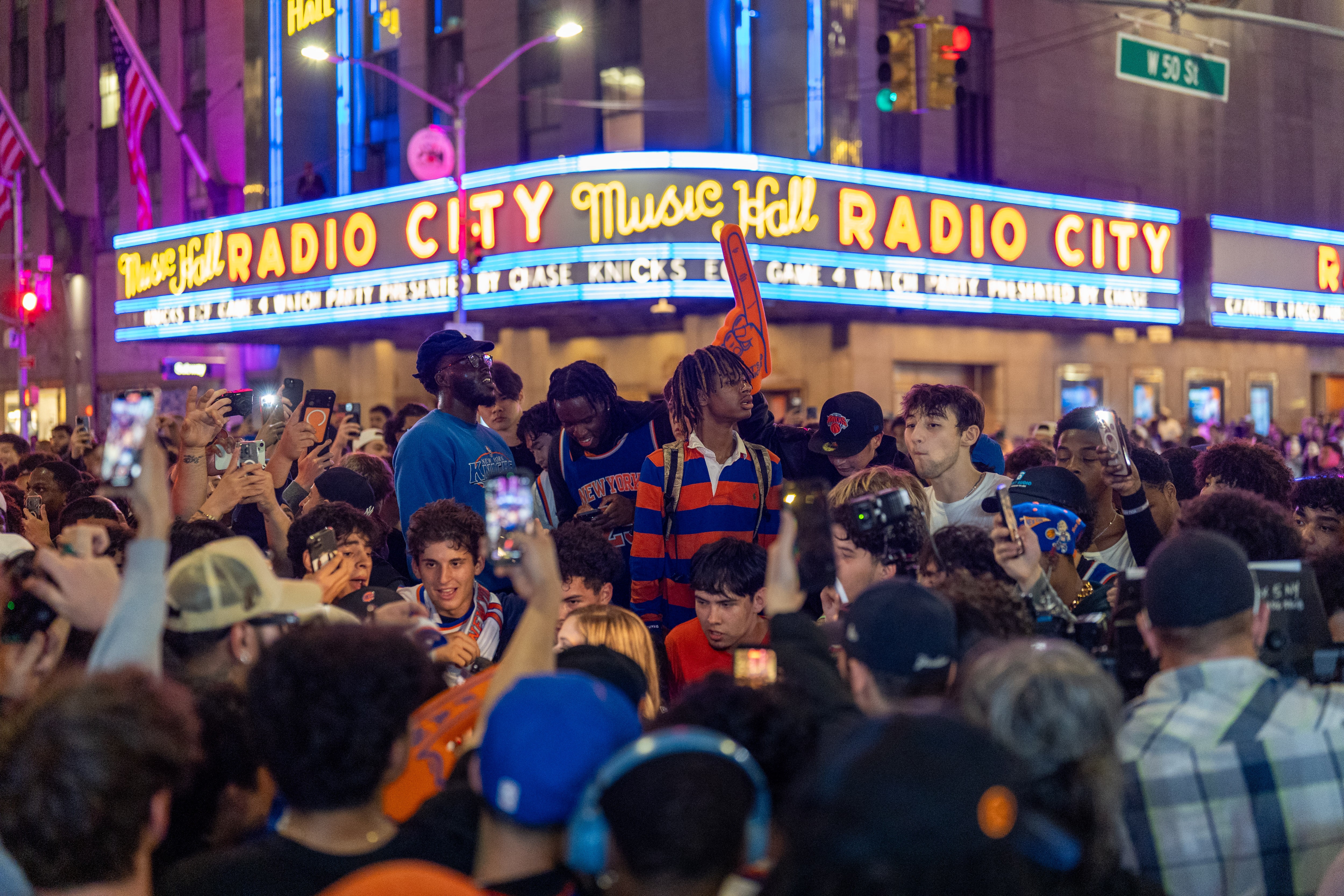 Multidão de jovens e adultos em frente ao Radio City Music Hall iluminado, muitos filmando com celulares, alguns usando bonés e camisas de basquete, em uma noite movimentada na cidade