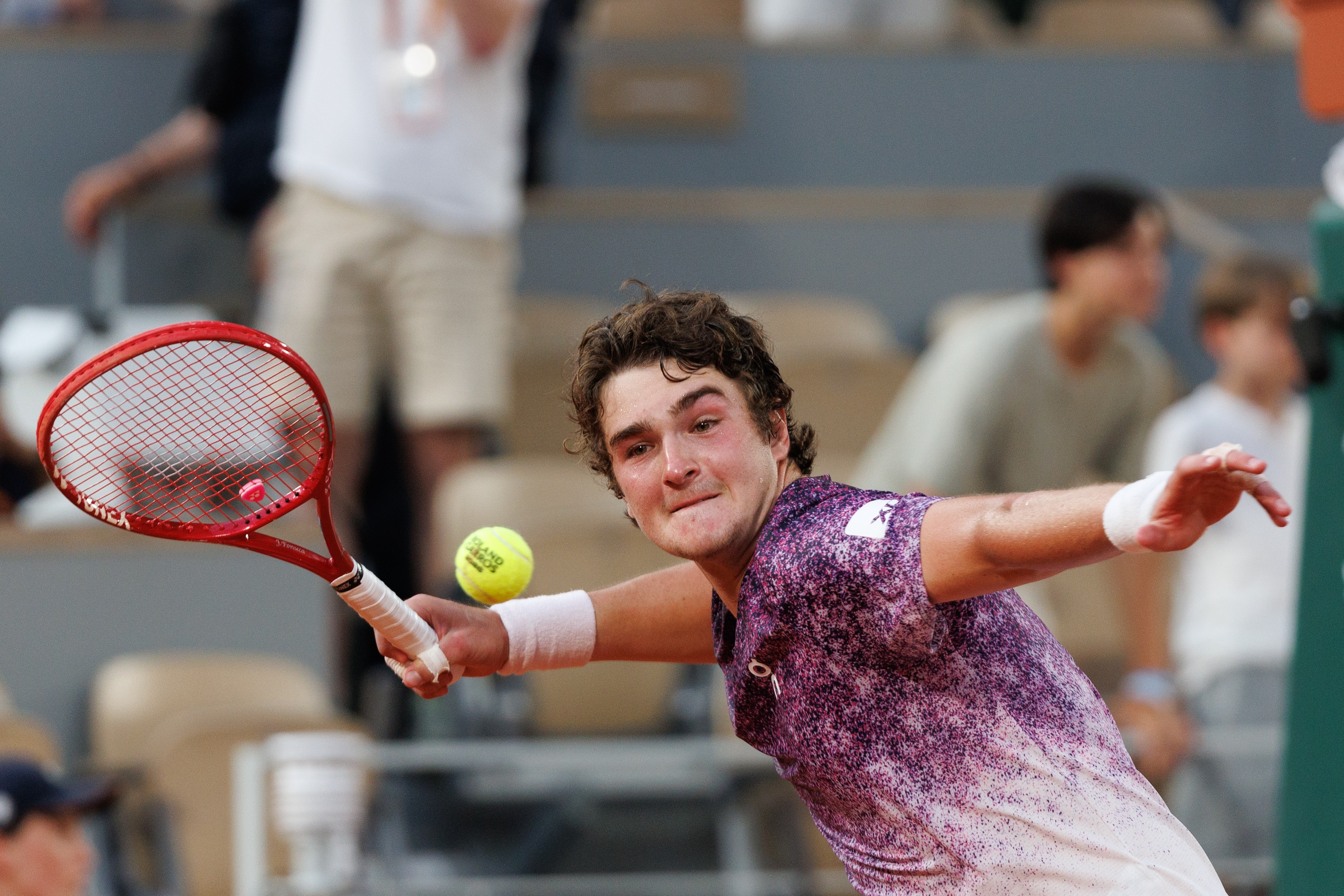 Jovem tenista com cabelo castanho e camisa roxa e branca, concentrado, rebatendo uma bola de tênis amarela com uma raquete vermelha em quadra de saibro. Ele usa uma munhequeira branca no braço direito. Pessoas desfocadas ao fundo.