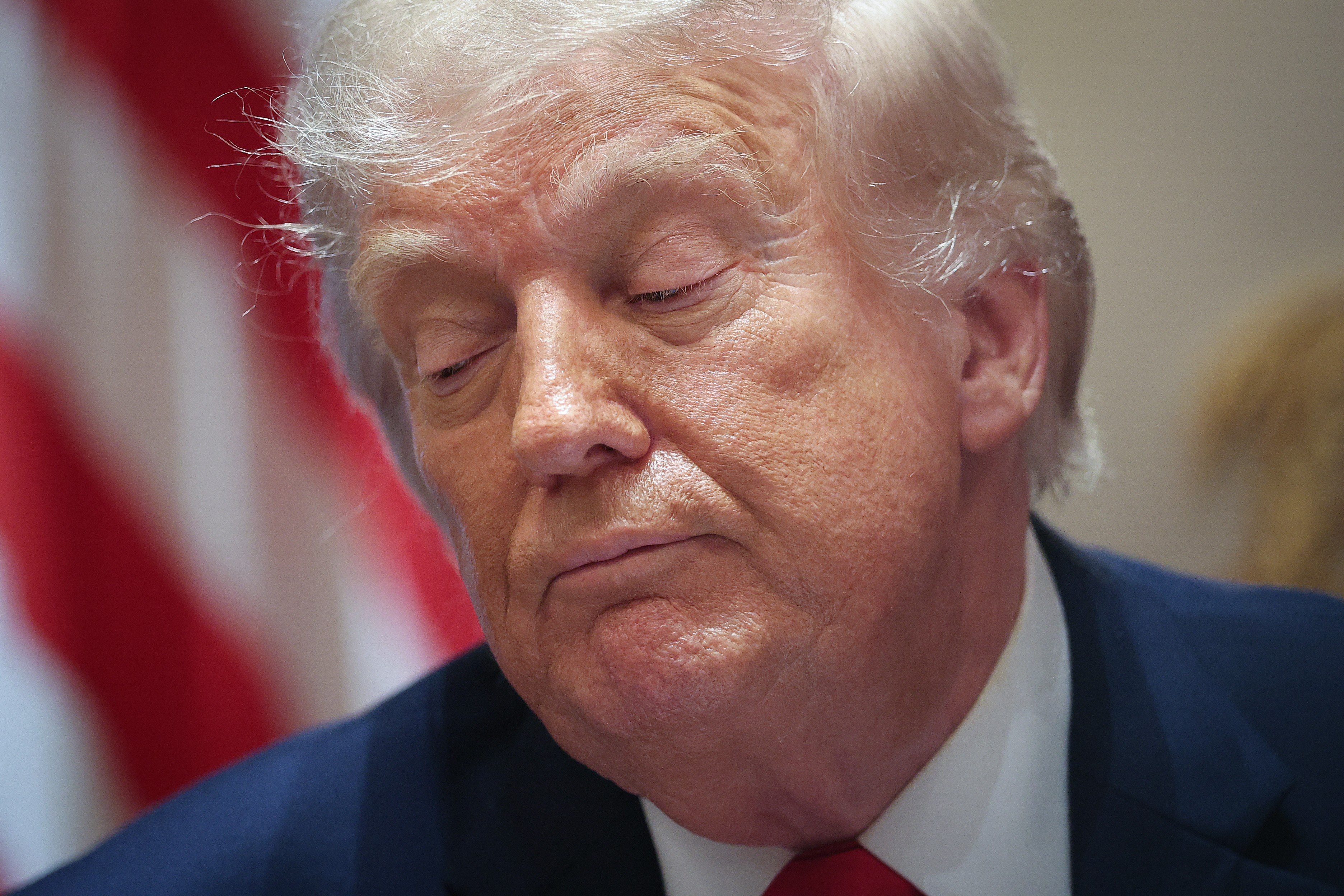 Donald Trump, com cabelos loiros e olhos fechados, usa terno azul marinho e gravata vermelha, com a bandeira dos EUA desfocada ao fundo