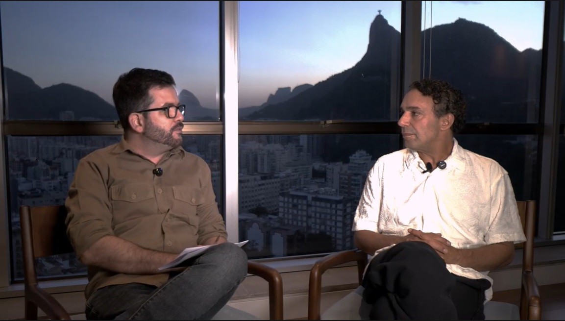 Dois homens sentados em cadeiras de madeira, um de camisa marrom e óculos, segurando papéis, e outro de camisa branca, com as mãos cruzadas. Ao fundo, uma janela exibe a paisagem noturna do Rio de Janeiro, com montanhas e edifícios, incluindo o Cristo Redentor no topo de uma montanha