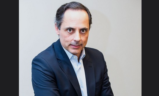 Homem de meia-idade, cabelos escuros e grisalhos, terno azul-marinho e camisa azul-clara, olhando diretamente para frente com expressão séria