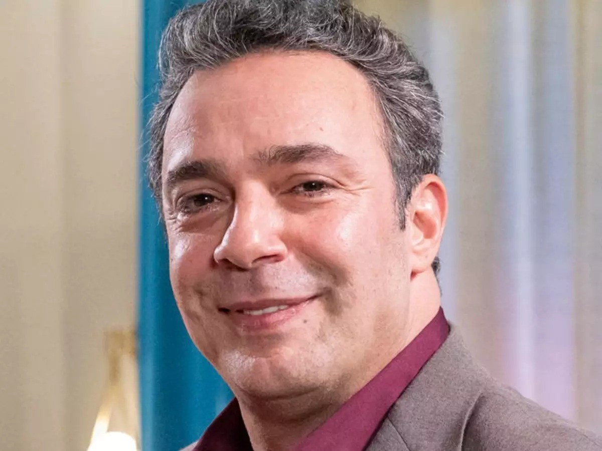 Homem de pele clara, cabelos grisalhos e curtos, sorrindo levemente, vestindo camisa vinho e blazer cinza, em ambiente interno com cortina azul ao fundo