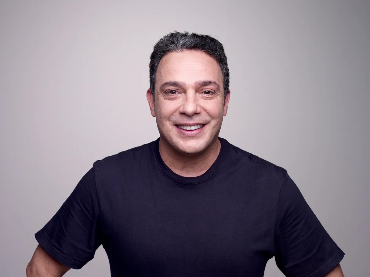 Homem de pele clara, cabelo escuro e grisalho, sorrindo com a boca aberta, vestindo camiseta preta, em fundo cinza claro