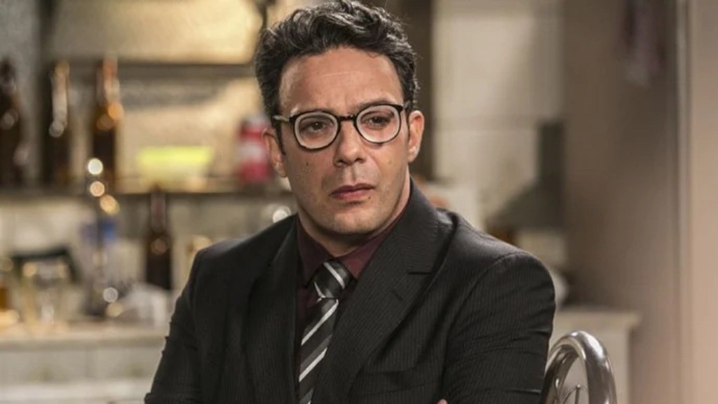 Homem de pele clara, cabelo escuro e óculos, vestindo terno preto, camisa vinho e gravata listrada, com expressão séria, olhando para frente. O fundo está desfocado, com garrafas e objetos em prateleiras