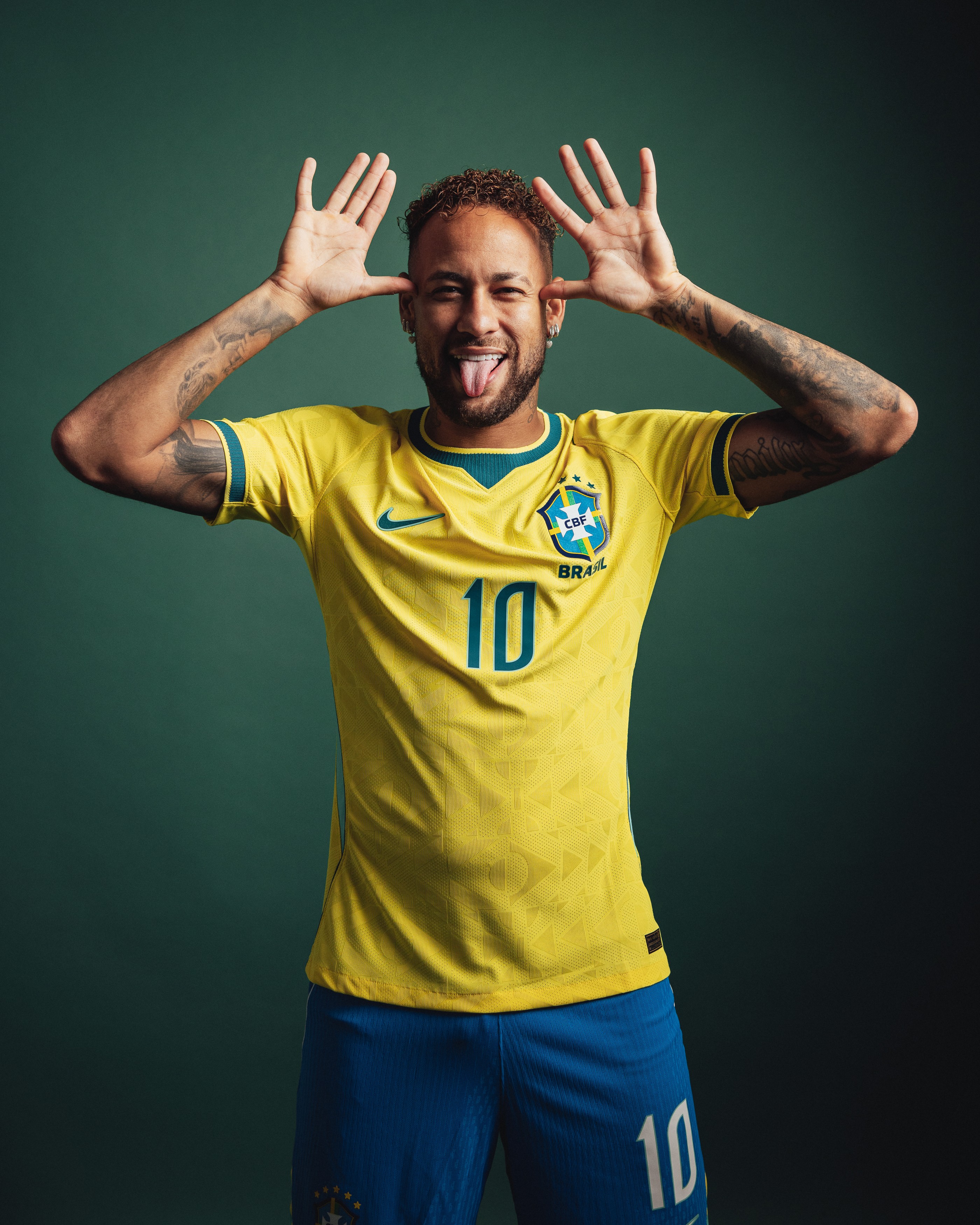 Neymar Jr. sorrindo e fazendo careta com a língua para fora, vestindo a camisa amarela da seleção brasileira com o número 10 e shorts azuis, em fundo verde escuro