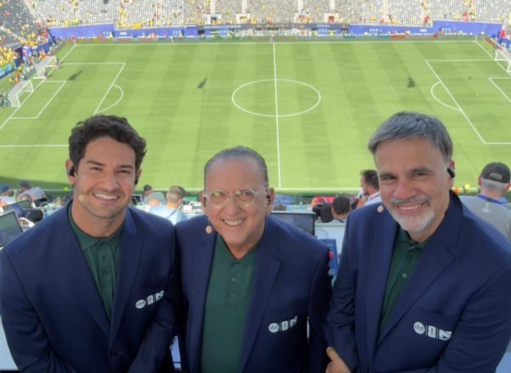Três homens sorridentes, de meia-idade, vestindo blazers azuis e camisas verdes, com fones de ouvido e microfones de lapela, posam em frente a um estádio de futebol com gramado verde e arquibancadas cheias