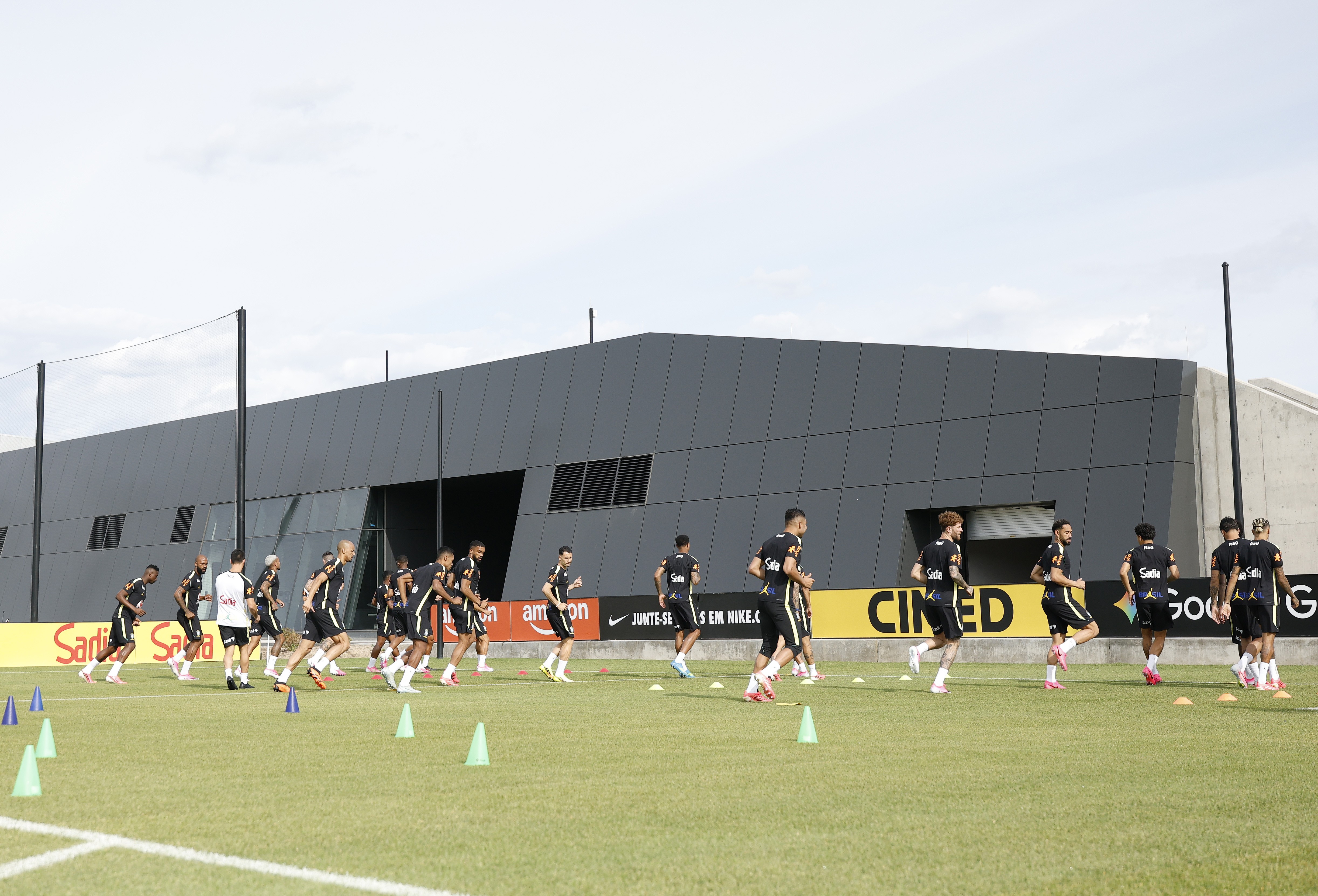 Jogadores de futebol, vestindo uniformes pretos, correm em fila em um campo de grama verde com cones de treino, em frente a um prédio cinza escuro e um banner de patrocinadores