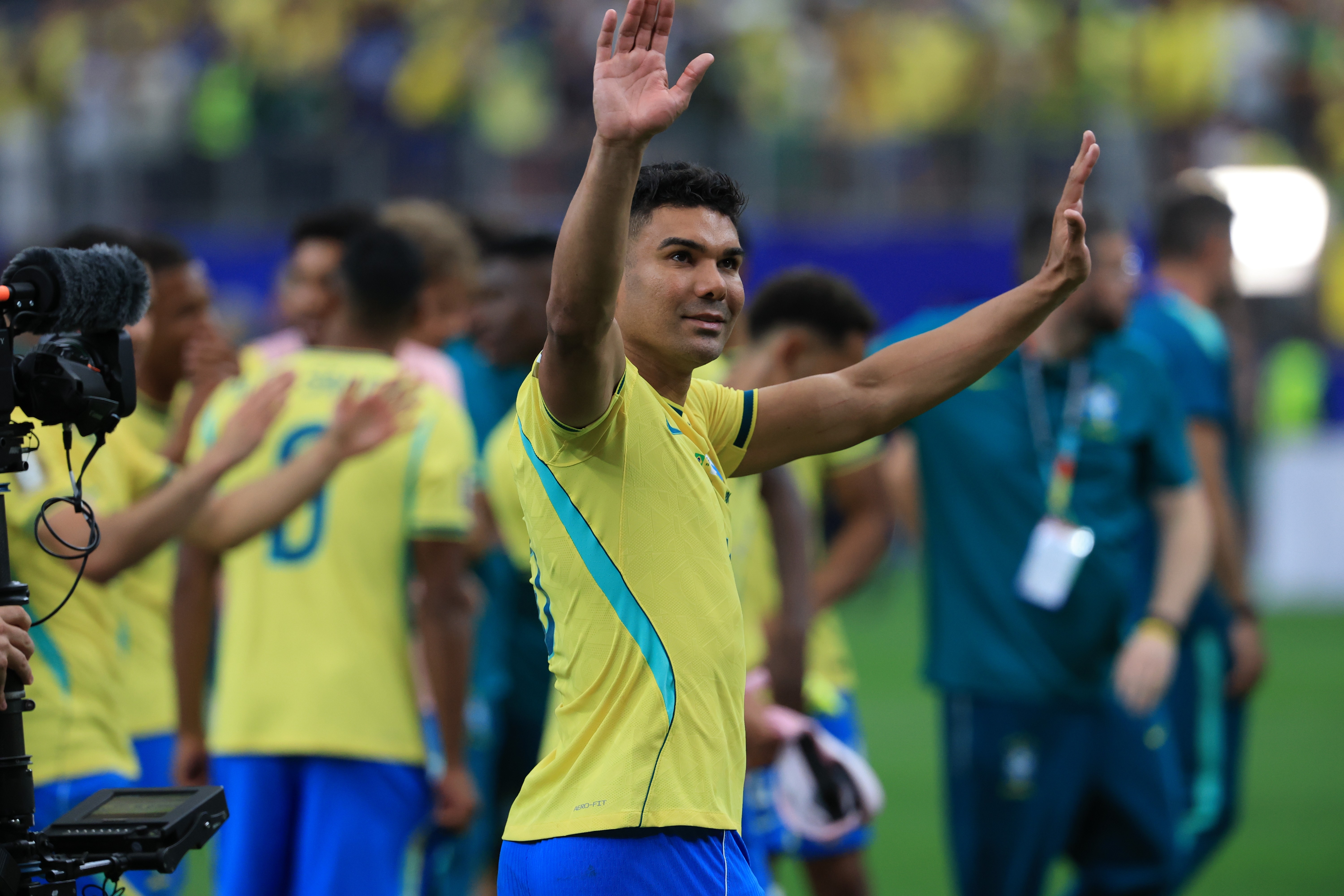Jogador de futebol Casemiro, com camisa amarela e detalhes azuis, sorri e acena com as duas m&atilde;os para a torcida em campo, com outros jogadores e equipe t&eacute;cnica ao fundo