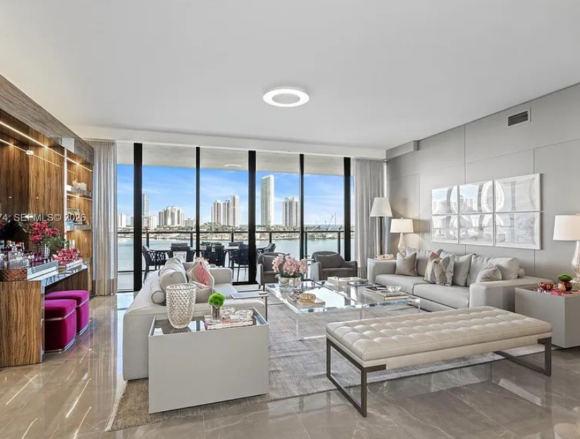 Sala de estar luxuosa com sofá cinza, mesa de centro de vidro, banco estofado bege e vista panorâmica para a cidade e o mar. À esquerda, um balcão de madeira com banquetas fúcsia e espelho.