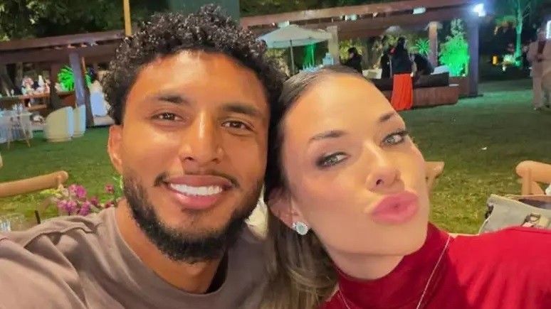Um homem sorridente com barba e cabelo cacheado escuro, vestindo camiseta cinza, ao lado de uma mulher com maquiagem e brincos, fazendo bico para a câmera. Ao fundo, um gramado verde com mesas e cadeiras iluminadas por luzes quentes e verdes, em um ambiente noturno de festa
