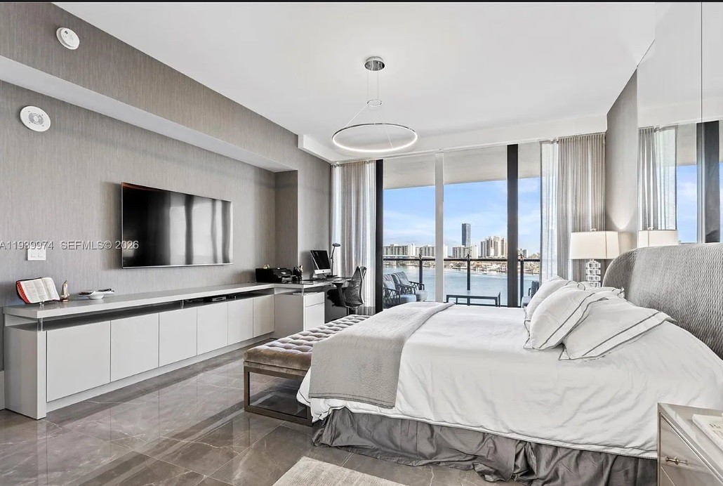 Quarto moderno com cama de casal branca, banco estofado cinza, armário baixo com televisão e escrivaninha. Uma janela ampla revela a vista para a cidade e o mar.
