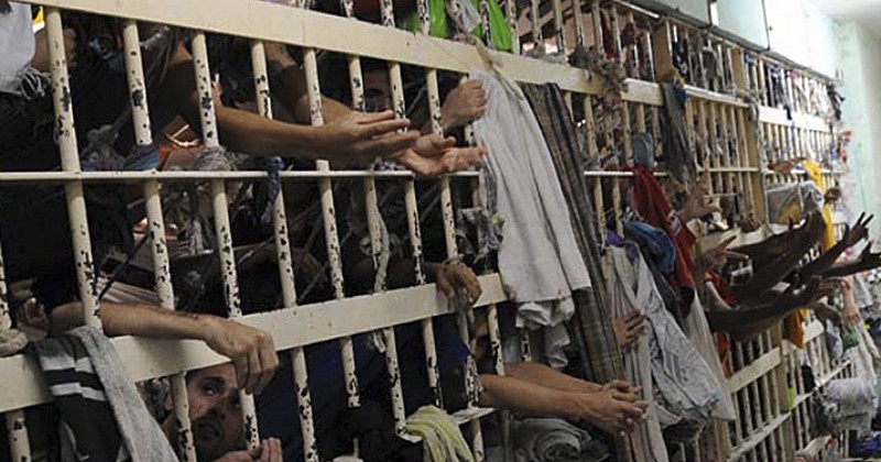 Muitos homens presos, com roupas simples, estendem as mãos e braços através das grades de celas superlotadas, que têm roupas penduradas, em um ambiente de prisão precário