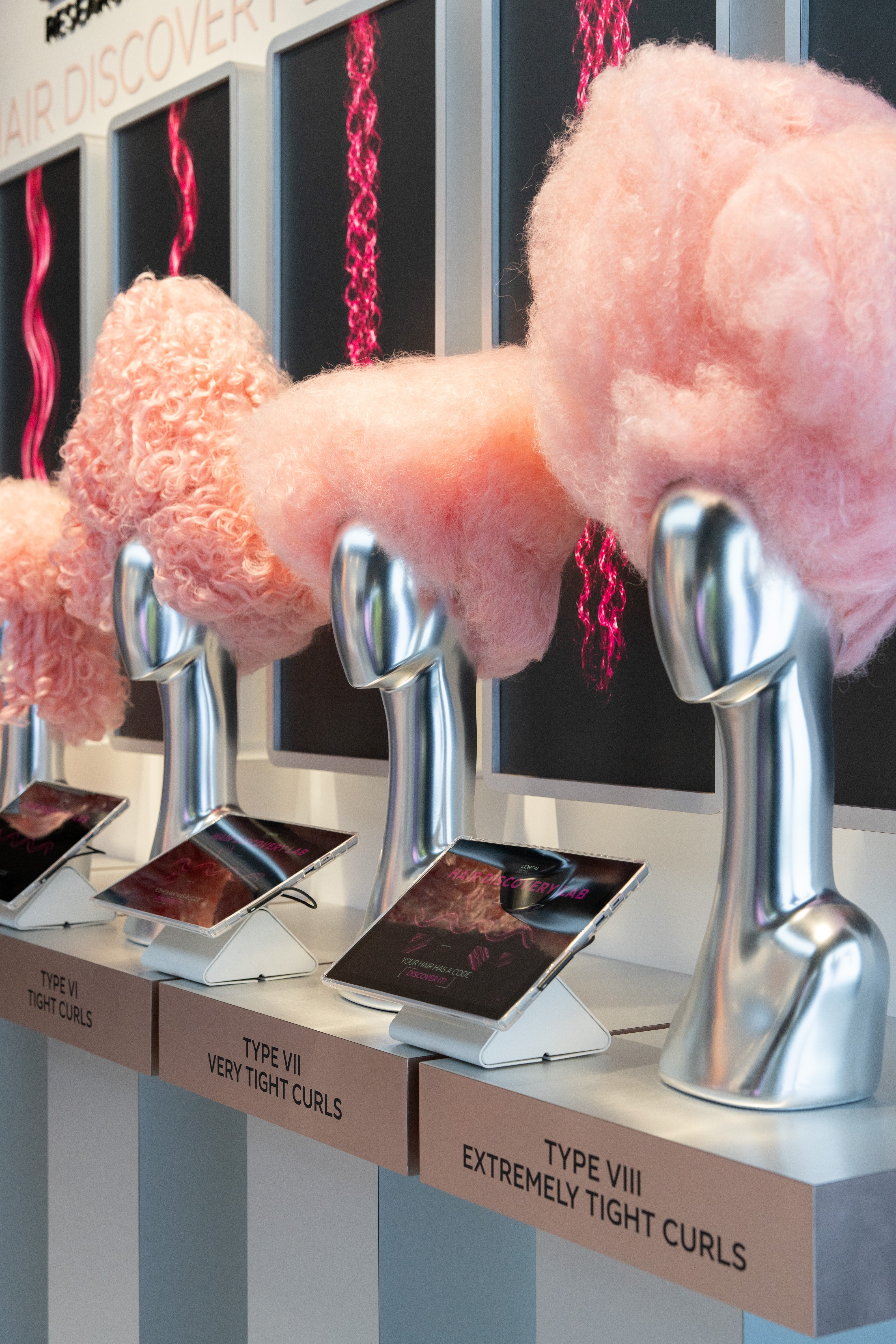 Exposição de tipos de cachos com manequins prateados exibindo perucas de algodão doce rosa e telas mostrando padrões de cabelo em neon rosa, com tablets e legendas TYPE VI TIGHT CURLS, TYPE VII VERY TIGHT CURLS e TYPE VIII EXTREMELY TIGHT CURLS
