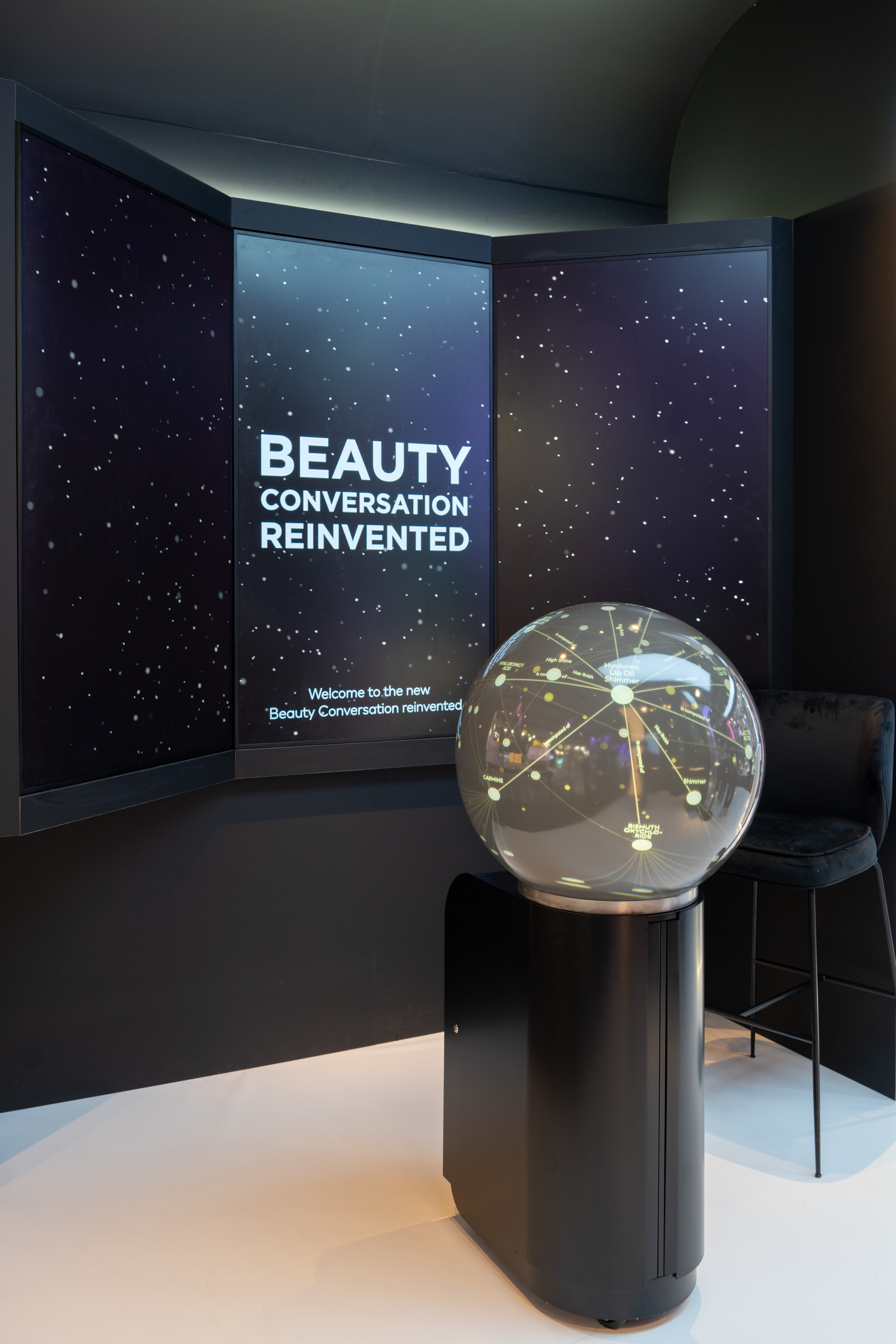 Ambiente escuro com telas exibindo um céu estrelado e o texto BEAUTY CONVERSATION REINVENTED. Uma esfera transparente com pontos luminosos e linhas interconectadas está sobre um pedestal preto, ao lado de um banco alto escuro