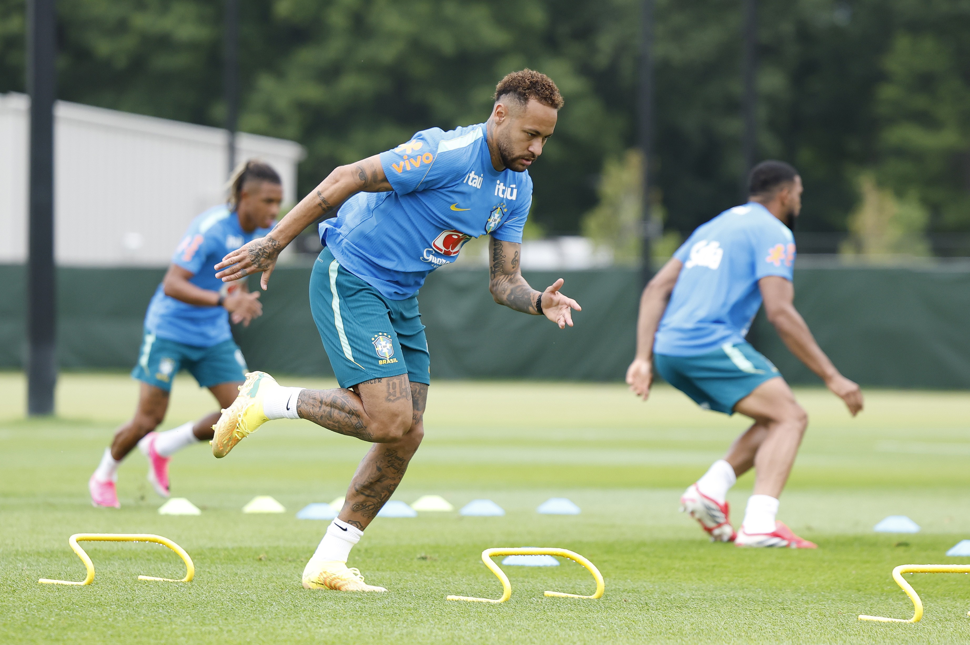 Neymar Jr. em primeiro plano, correndo agachado para a direita, usando camisa azul e shorts verde-escuro da seleção brasileira, com chuteiras amarelas. Outros dois jogadores, um com cabelo trançado e outro de costas, também correm no campo verde, com pequenos obstáculos amarelos no chão