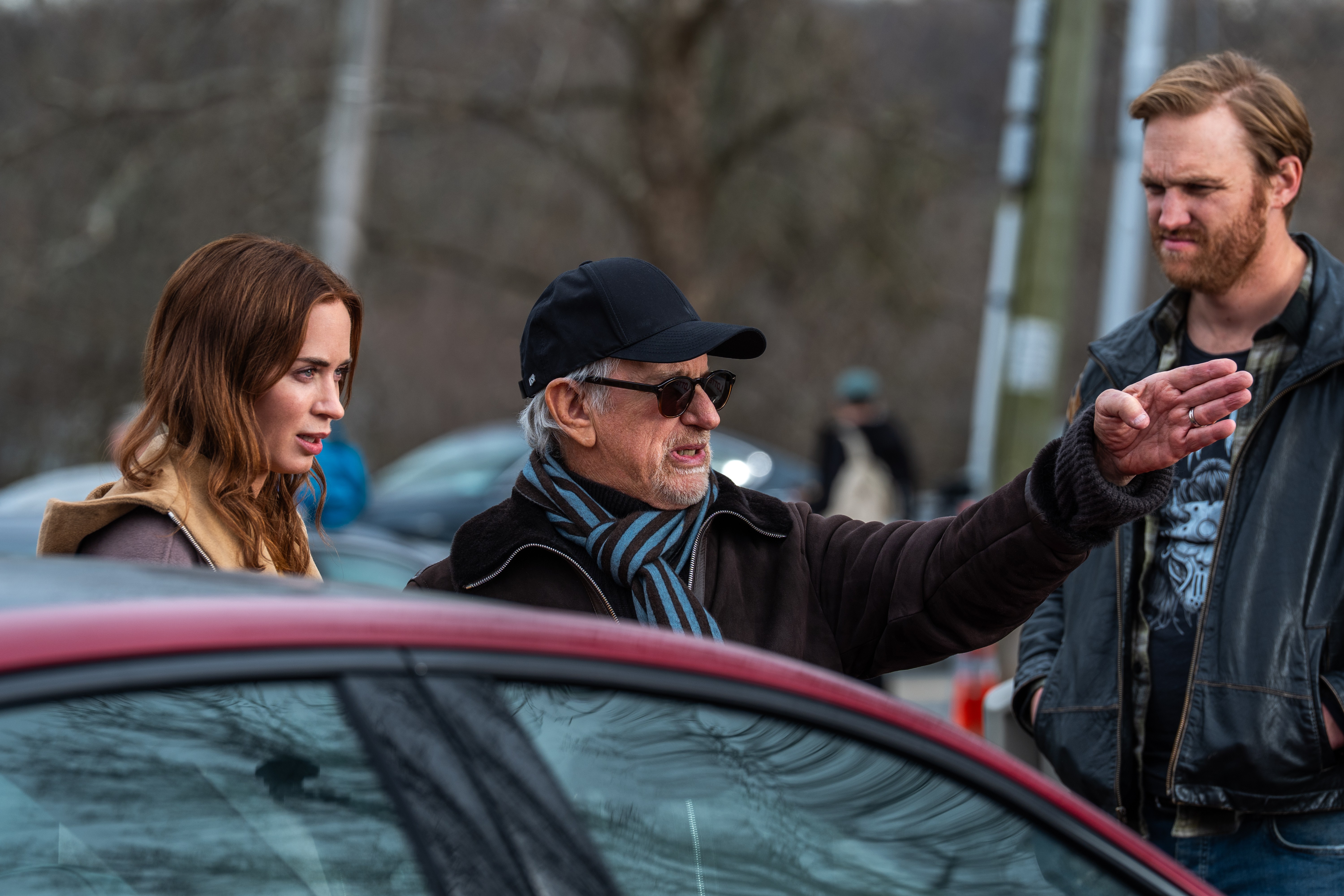 O diretor Steven Spielberg, de boné e óculos escuros, gesticula com a mão direita enquanto conversa com a atriz Emily Blunt e o ator Wyatt Russell, em um cenário externo com carros e árvores ao fundo