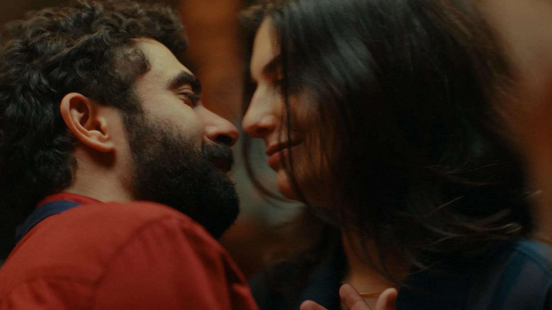 Um homem barbudo de cabelo encaracolado e uma mulher de cabelo escuro se aproximam para um beijo, com sorrisos sutis nos lábios. Ele veste uma camisa vermelha, ela uma blusa escura com um colar dourado. A iluminação é suave e quente