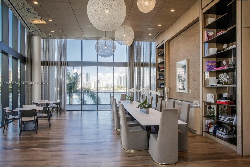 Sala de jantar moderna com mesa longa e cadeiras estofadas cinzas, sob grandes luminárias esféricas. Ao fundo, janelas do chão ao teto revelam vista para o mar e prédios urbanos. Estantes com livros e objetos decorativos adornam a parede lateral.