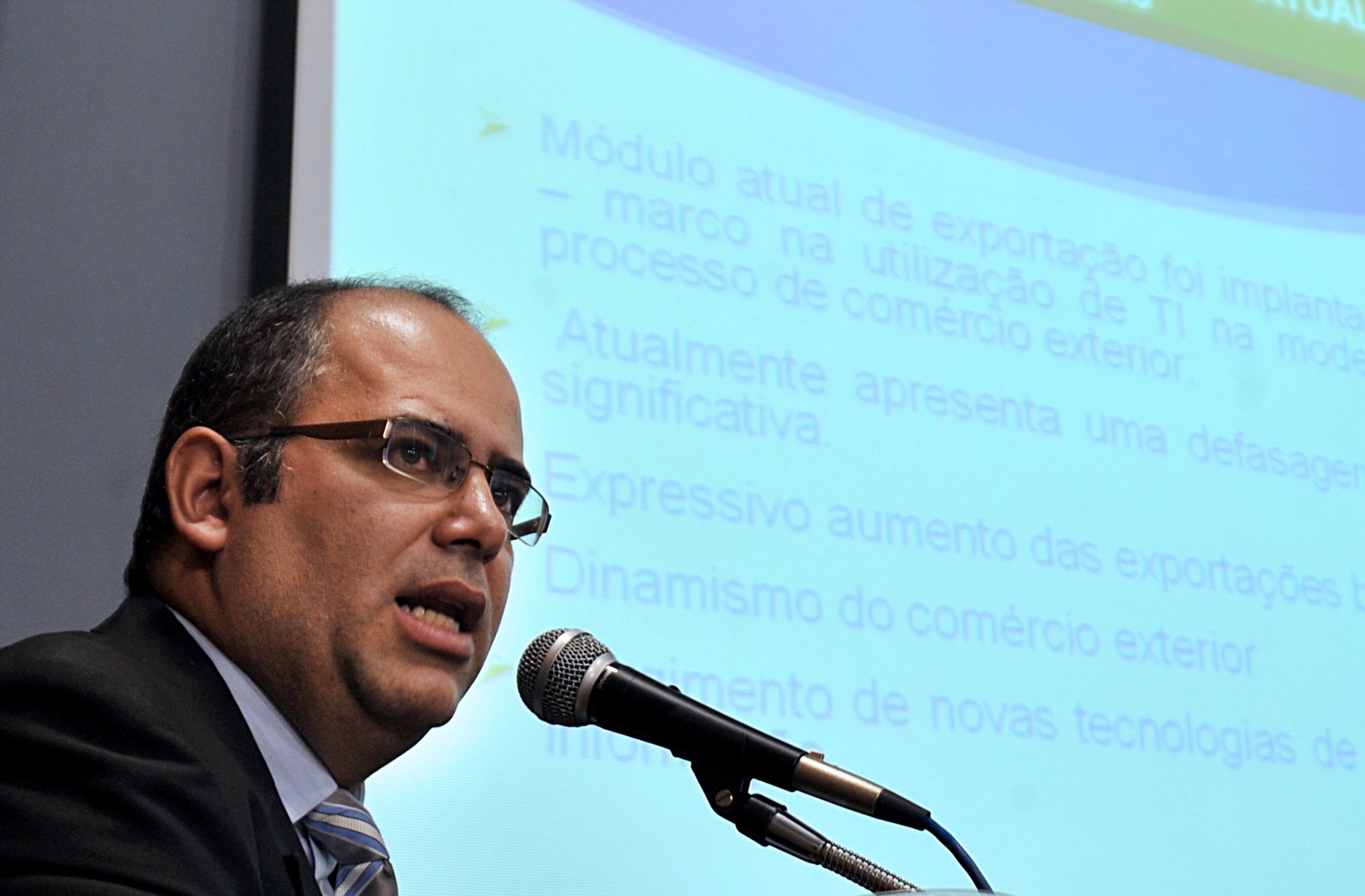 Homem calvo de óculos e terno escuro, gravata listrada, falando em um microfone, com uma projeção azul de slides ao fundo