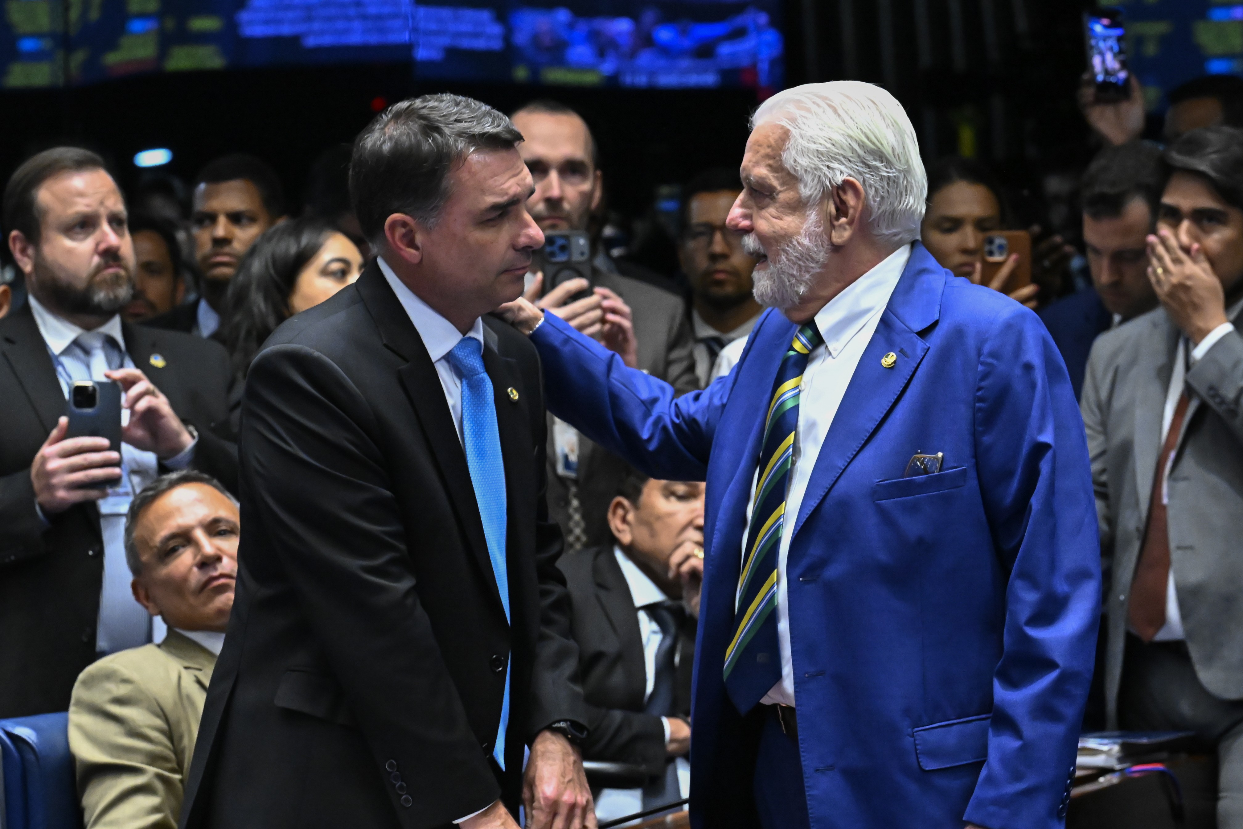 Dois homens brancos, um de terno azul e barba branca, e outro de terno preto e gravata azul, conversam em meio a uma multidão em um ambiente interno. O homem de azul toca o ombro do outro, que o olha atentamente.