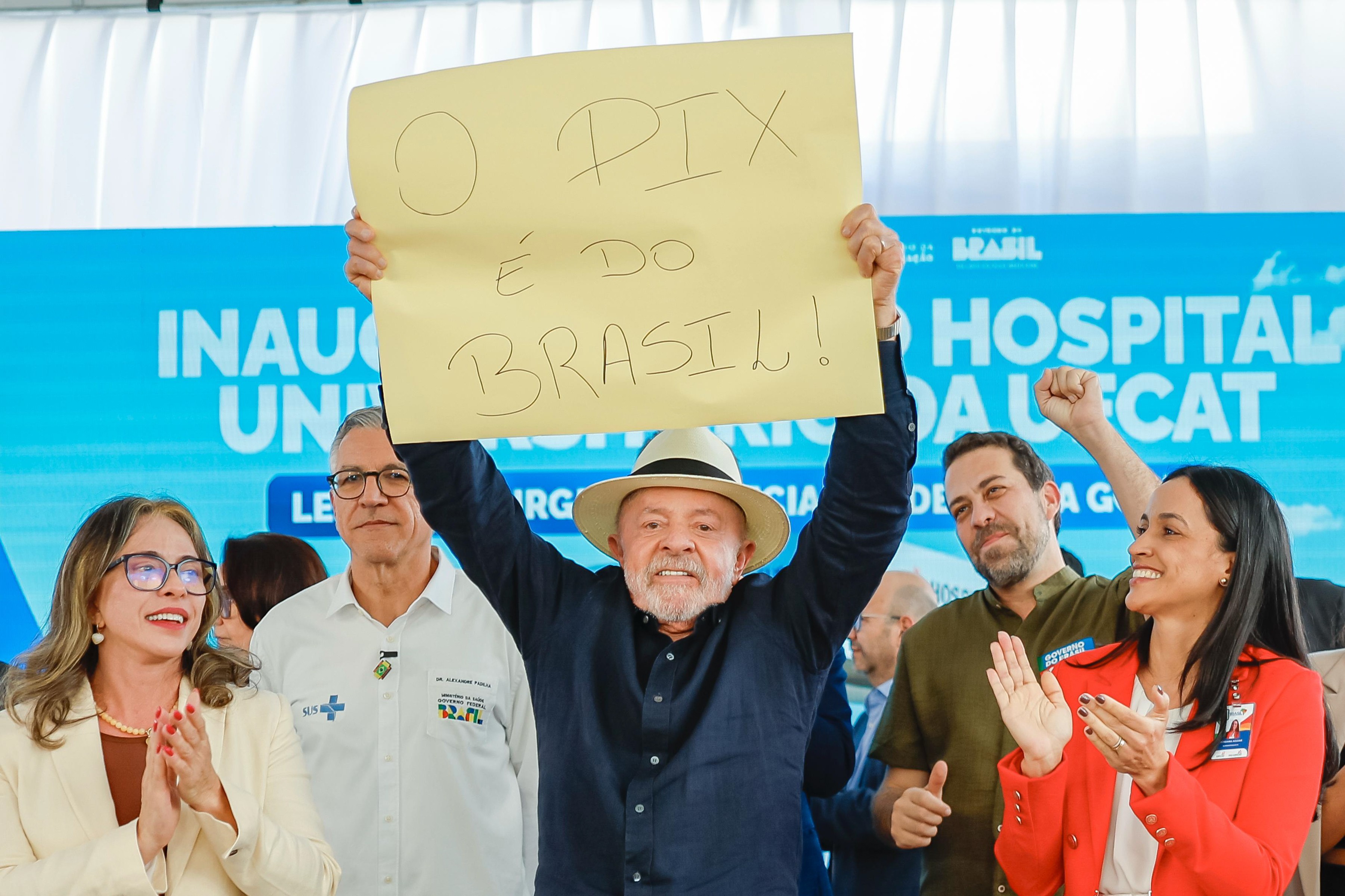 Lula, sorrindo e usando chapéu de palha, ergue um cartaz amarelo com "O PIX É DO BRASIL!". Ao seu lado, pessoas aplaudem e celebram, com um banner azul de "INAUGURAÇÃO HOSPITAL" ao fundo