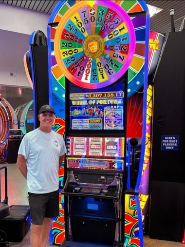 Homem sorridente de boné preto, camiseta branca e bermuda escura posa ao lado de uma máquina caça-níqueis colorida com uma grande roda da fortuna no topo, em um cassino