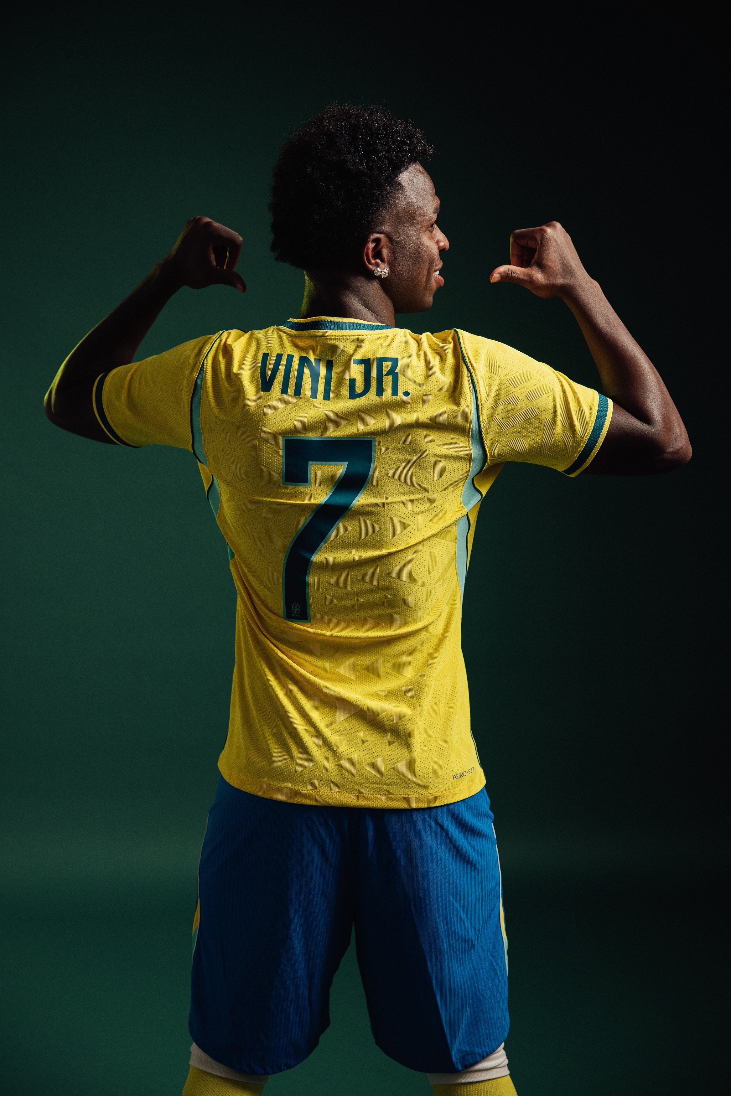 Jogador de futebol Vinicius Jr. de costas, vestindo camisa amarela da seleção brasileira com VINI JR. e o número 7 em azul, e shorts azuis. Ele flexiona os braços, mostrando os bíceps, e olha para a direita, em um fundo verde escuro