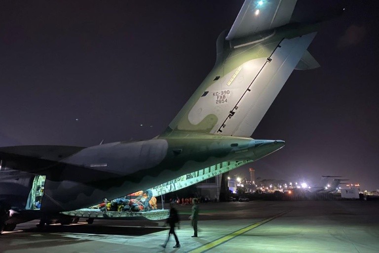 Avião militar KC-390 da FAB, camuflado em tons de verde e cinza, com a rampa traseira aberta e carga visível, estacionado em pátio de aeroporto à noite. Duas pessoas, uma em movimento e outra parada, estão próximas à aeronave. Ao fundo, luzes de outros aviões e edifícios do aeroporto iluminam a escuridão