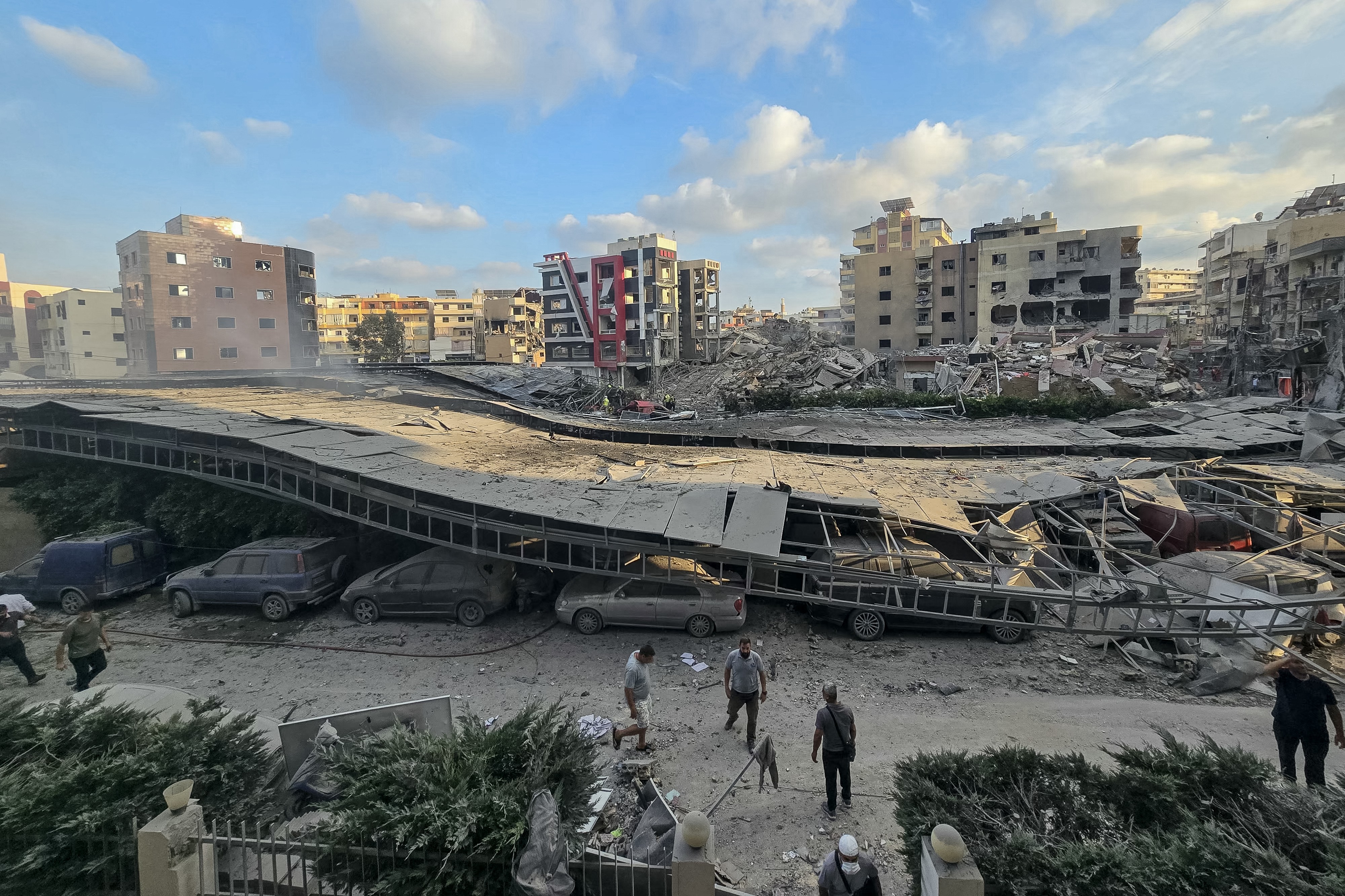 Vista aérea de uma rua com um viaduto parcialmente desabado, esmagando carros embaixo. Ao fundo, edifícios destruídos e escombros. Pessoas caminham pela rua, avaliando os danos sob um céu azul com nuvens esparsas