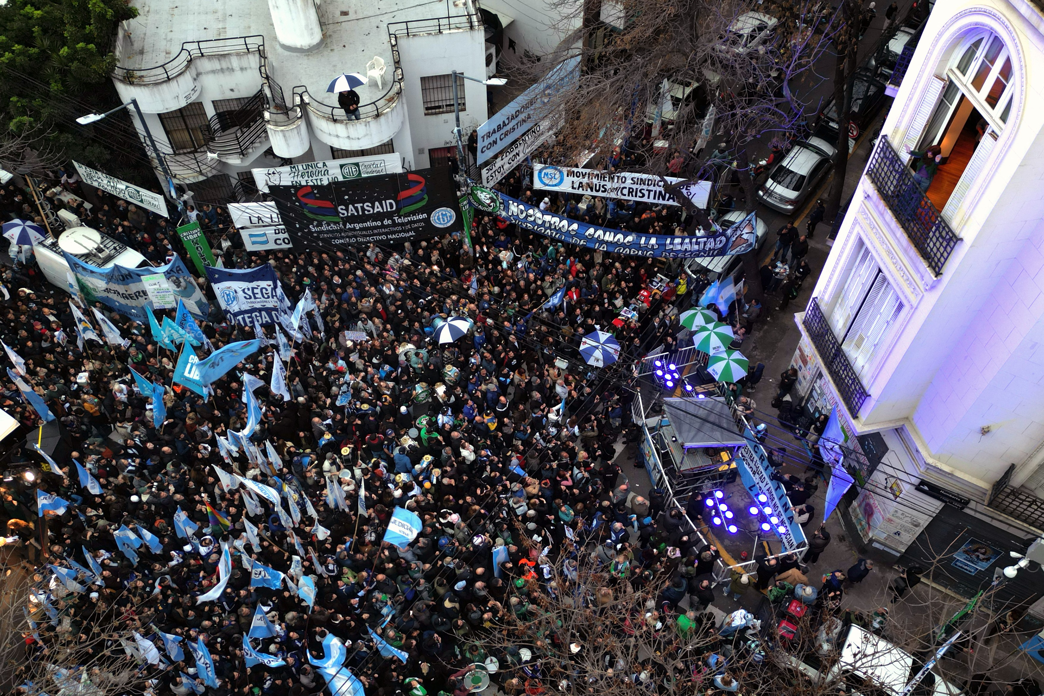 Multidão de pessoas em uma rua, com muitas bandeiras azuis e brancas, algumas com o sol da bandeira argentina. Há faixas penduradas em prédios, uma delas com SATSAID e outra com MOVIMIENTO SINDICAL LANUS CRISTINA. Guarda-chuvas abertos em azul e branco, e verde e branco, espalhados pela multidão. Um palco com luzes azuis está à direita, e um prédio branco com sacadas e janelas altas aparece no canto superior direito. Carros estacionados e árvores secas completam o cenário urbano Former Argentine president (2007–2015) Cristina Kirchner appears in the balcony of her home as she remains under house arrest in Buenos Aires on June 10, 2026. Argentine courts have issued a warning to former President Cristina Kirchner (2007–2015) as she marked the first of her six years of house arrest for corruption on June 17, 2026, due to the displays of support she is receiving outside her home. The courts reminded her that they may revoke her house arrest if she breaches the conditions set out by “disturbing the peace of the neighbourhood”, after hundreds of people gathered outside her home under the slogan “Free Cristina” on June 14, 2026.